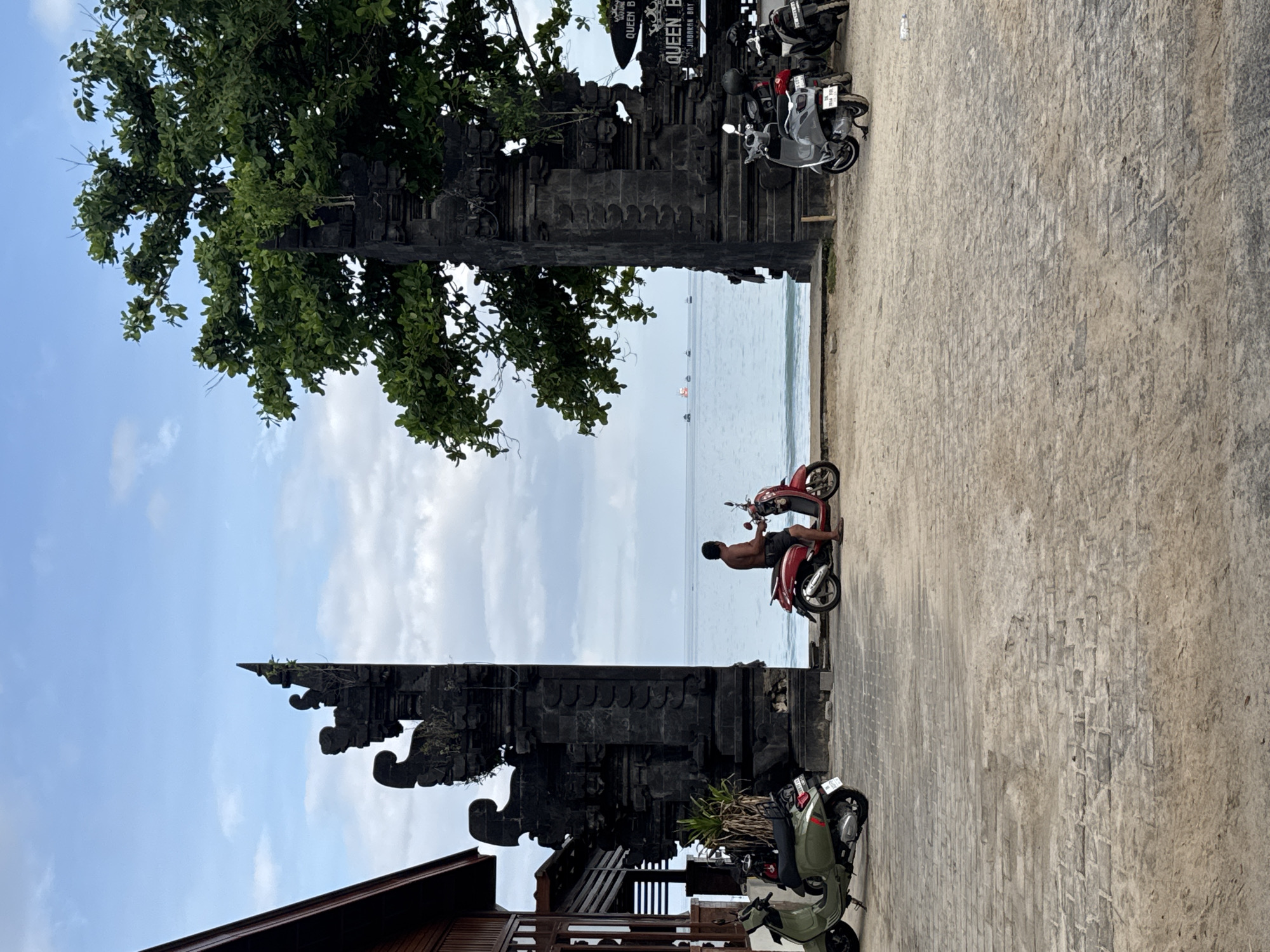 The image depicts a coastal scene in Kuta Selatan, Indonesia. A traditional Balinese gate frames a view of the ocean and beach. A man sits on a red scooter near the beach's edge, facing the water. Several parked motorbikes are visible on the right side of the gate and on the left side. A tree stands to the left of the gate, its foliage partially obscuring the sky. The sky is partly cloudy. The ground is paved.
