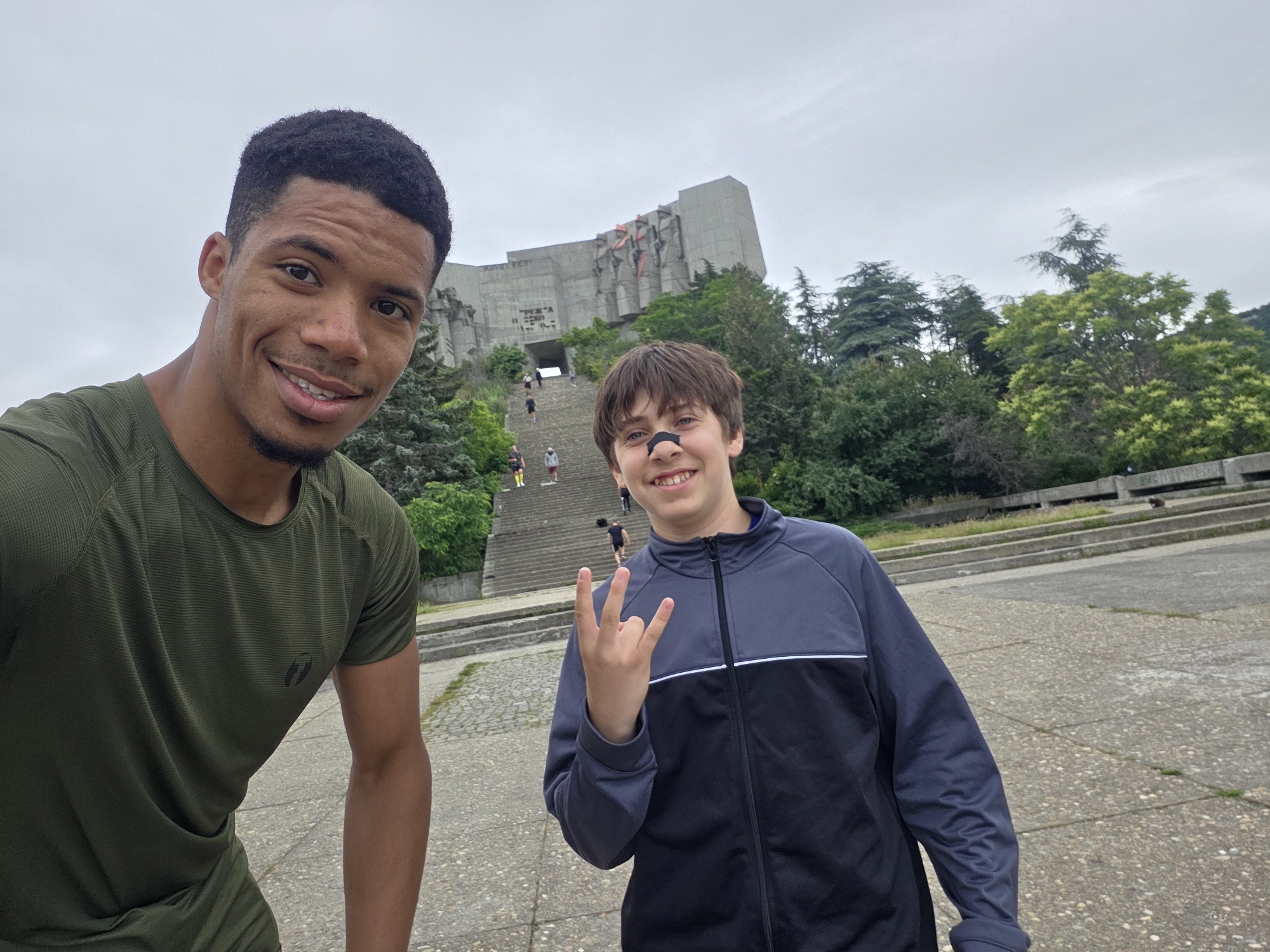 Two young men are taking a selfie outdoors in front of a large monument. The man on the left, facing the camera directly, is wearing a dark green t-shirt. The man on the right, slightly to the right of the frame, is wearing a dark blue and gray zip-up jacket and making a peace sign with his right hand. He has a black bandage on his nose. Behind them, a wide concrete staircase leads up to a massive, Brutalist-style concrete monument with geometric carvings and red accents. Trees and vegetation surround the monument and stairs. The sky is overcast and gray. The monument is the Monument to the Bulgarian-Soviet Friendship in Varna, Bulgaria.