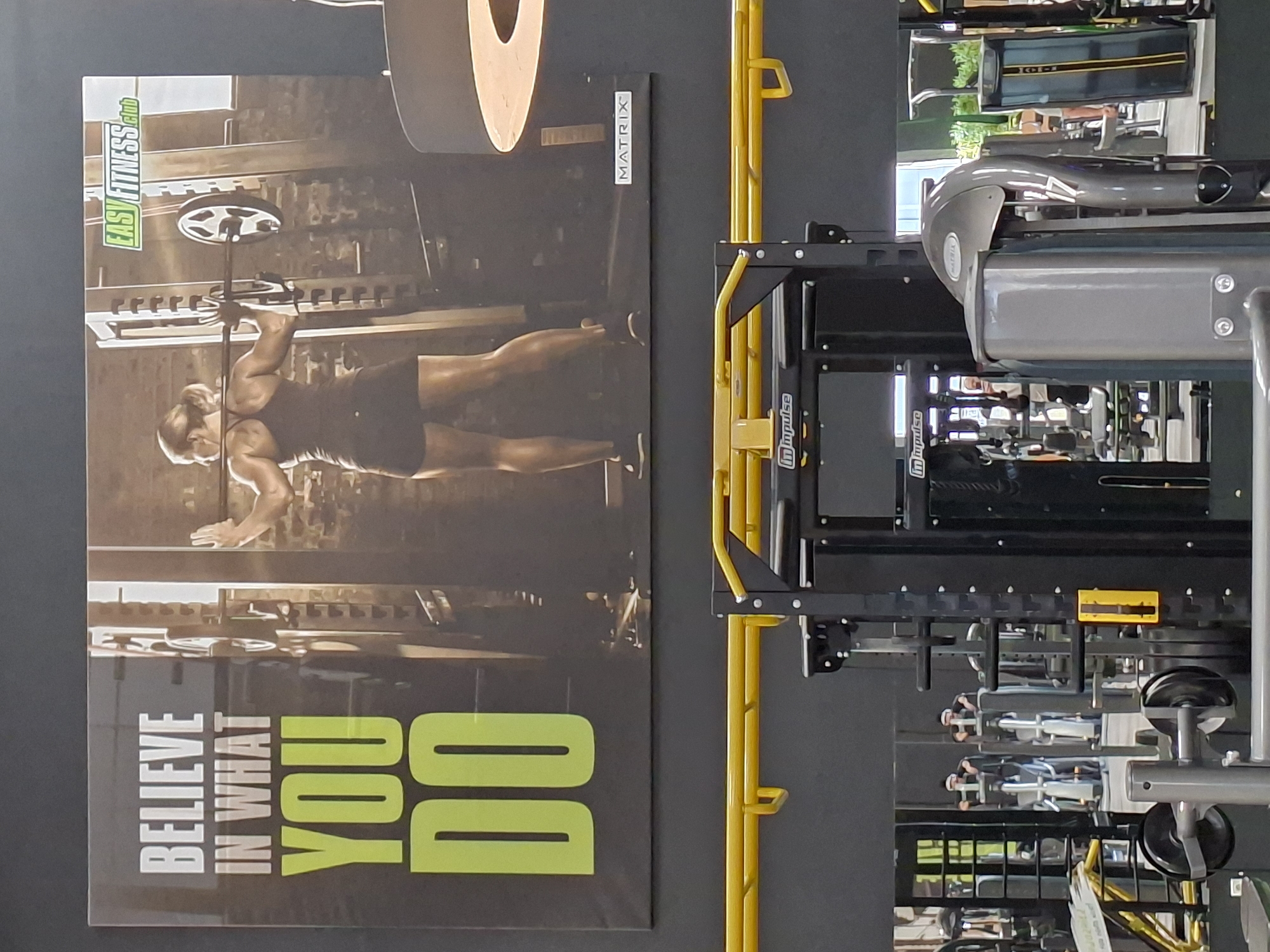 The image depicts the interior of a modern fitness center, likely located in Baden-Baden, Germany, given the context. The primary focus is a large motivational poster on the wall featuring a woman performing a squat with a barbell. The poster has bold text that reads "BELIEVE IN WHAT YOU DO" in white and neon green. Above the woman's head, the logo "EASY FITNESS Club" is visible. To the right of the poster, the "MATRIX" brand logo is displayed.
Below the poster, a gym environment is evident with various exercise equipment. A black and yellow pull-up bar or rig is prominent in the foreground, along with weight racks and other machinery. Reflections in the mirrored walls reveal more gym equipment and possibly other patrons exercising, though they are not clearly identifiable. The overall lighting suggests an indoor setting with artificial illumination. The color palette is dominated by dark grays, blacks, and accents of yellow and neon green.