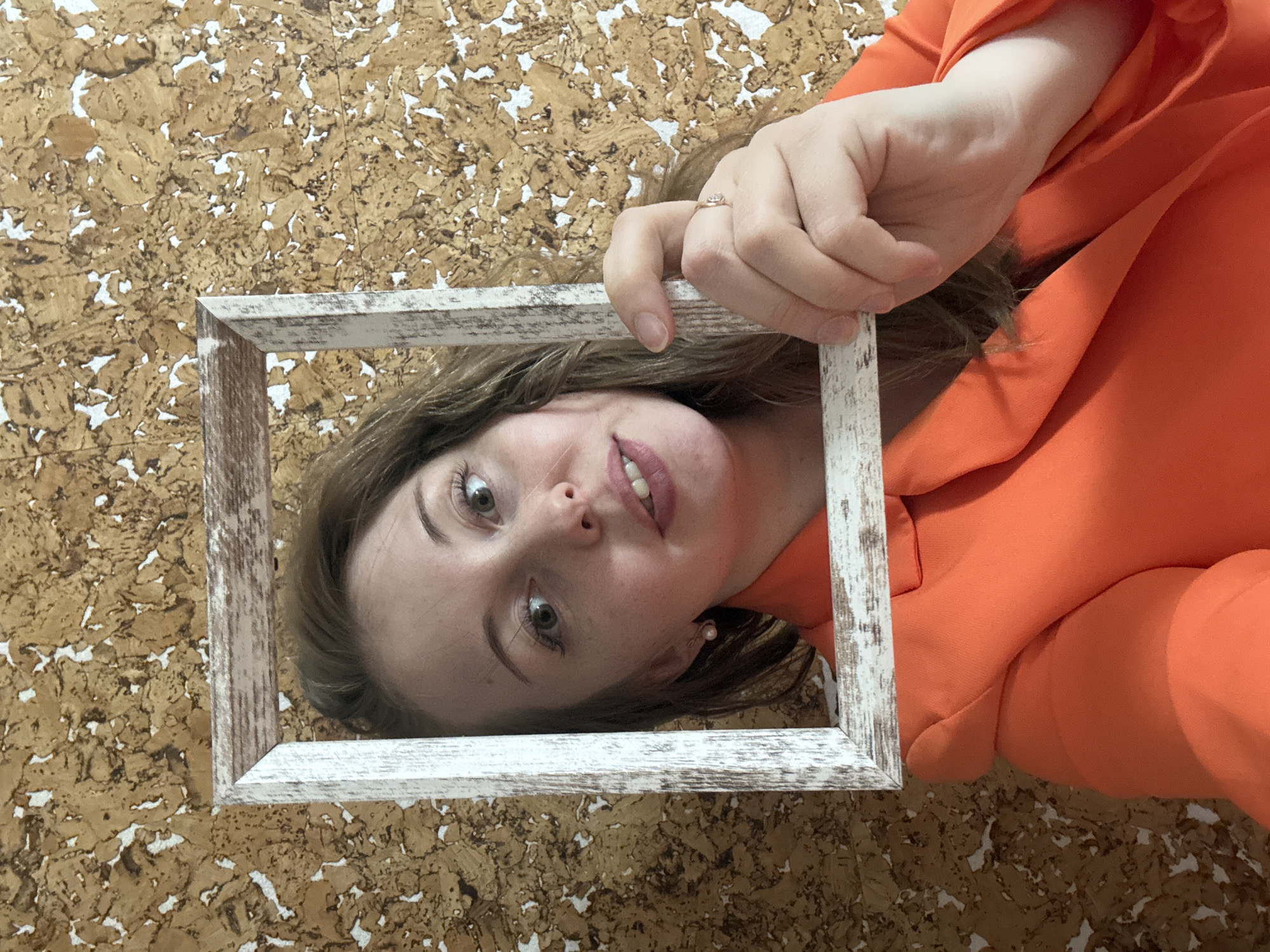A woman in an orange jacket holds a white frame around her face. The background is a textured, light-brown and white surface. The location is not discernible from the provided image.