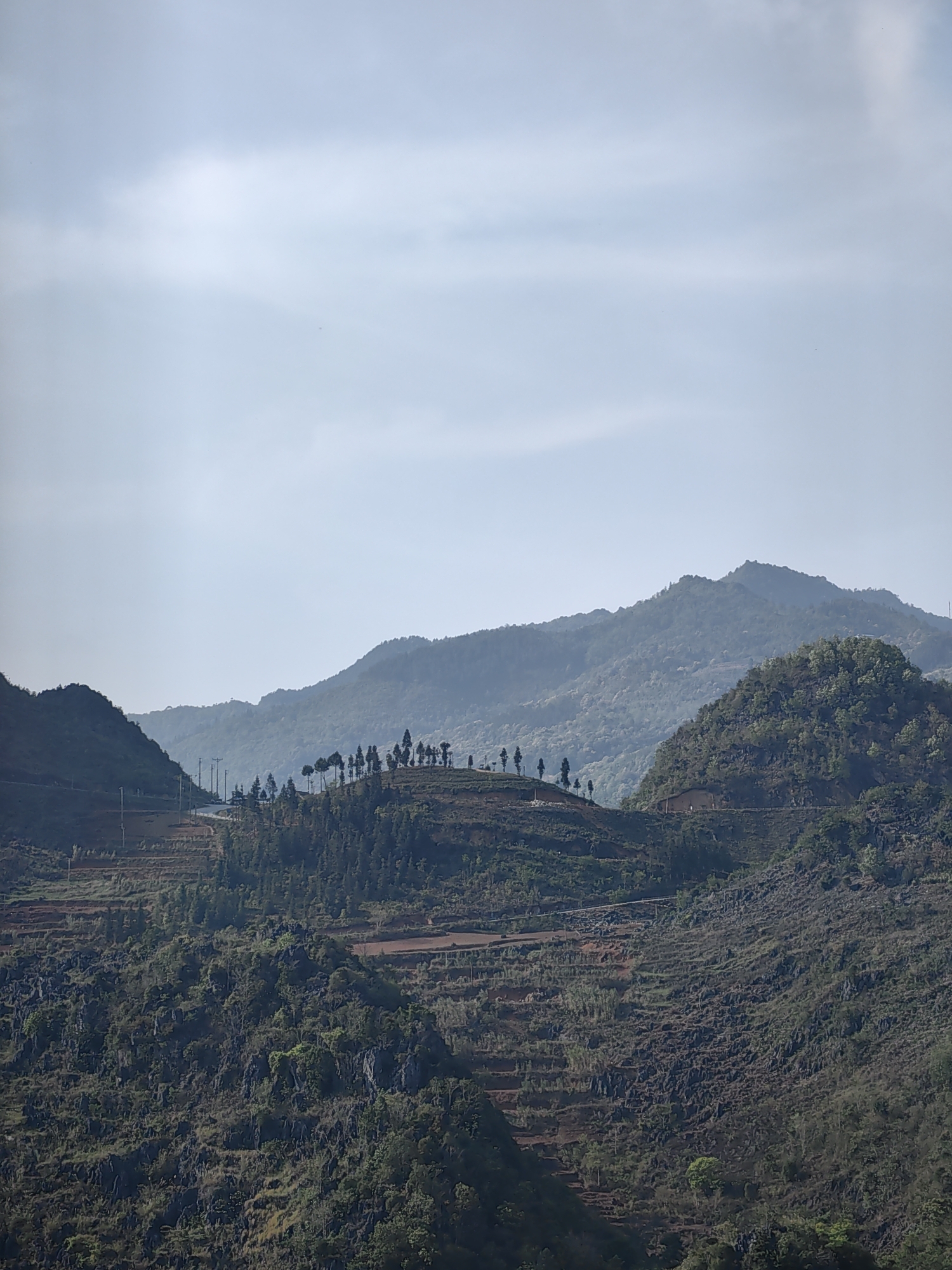This is a photograph taken in the Đồng Văn Commune, Vietnam, showing a landscape of layered mountains. The foreground is composed of rocky terrain interspersed with greenery. A road is visible, traversing the mountain's slope, with terraced fields carved into the hillside. Further up, another mountain features a cluster of trees atop its peak, with power lines running alongside the road. The background consists of more distant, hazy mountains under a light, overcast sky.