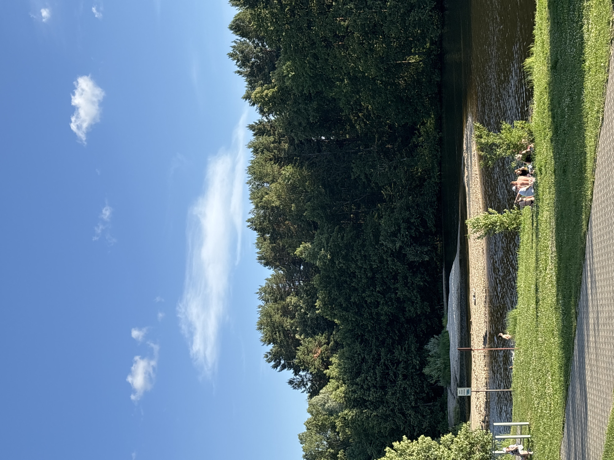 The image captures a sunny day with a clear blue sky and sparse clouds. The scene depicts a lakeside or riverside area with a dense line of lush green trees bordering a body of water. On the sandy bank, several people are visible, some sitting and relaxing, and others potentially wading in the shallow water. The water itself appears calm and reflects the light. To the right of the water, a well-maintained grassy area leads to a paved pathway. A sign is partially visible near the water's edge, but the text is not legible. Further to the right, a white bench is placed on the grass. The overall atmosphere suggests a leisurely day spent outdoors, possibly at a park or recreational area. The time of day appears to be late morning or early afternoon, given the bright sunlight and clear conditions.