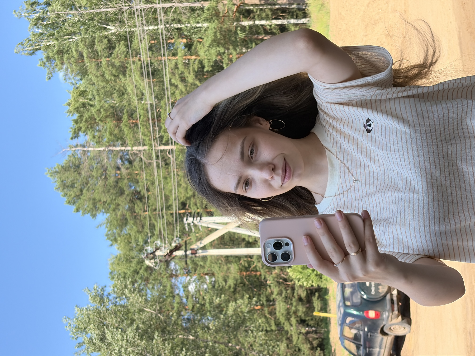 A young woman is taking a selfie outdoors. She is holding a light pink smartphone in her left hand, angled towards herself. She has her right hand in her hair, on the right side of her head. She is wearing a light-colored striped t-shirt with a small embroidered figure on it. She has hoop earrings and two rings on her left hand. Her hair is long and brown, flowing past her shoulders.
The background shows a clear blue sky with some scattered clouds. There are tall green pine trees and some birch trees. A power line pole with wires runs horizontally across the mid-ground. To the left of the woman, a dark green SUV is parked on a dirt road. The ground appears to be sandy. The overall setting suggests a rural or suburban area. The image is well-lit, indicating it was taken during the daytime, likely in the summer or early fall due to the lush greenery. The image was taken in Noviy, Russia.