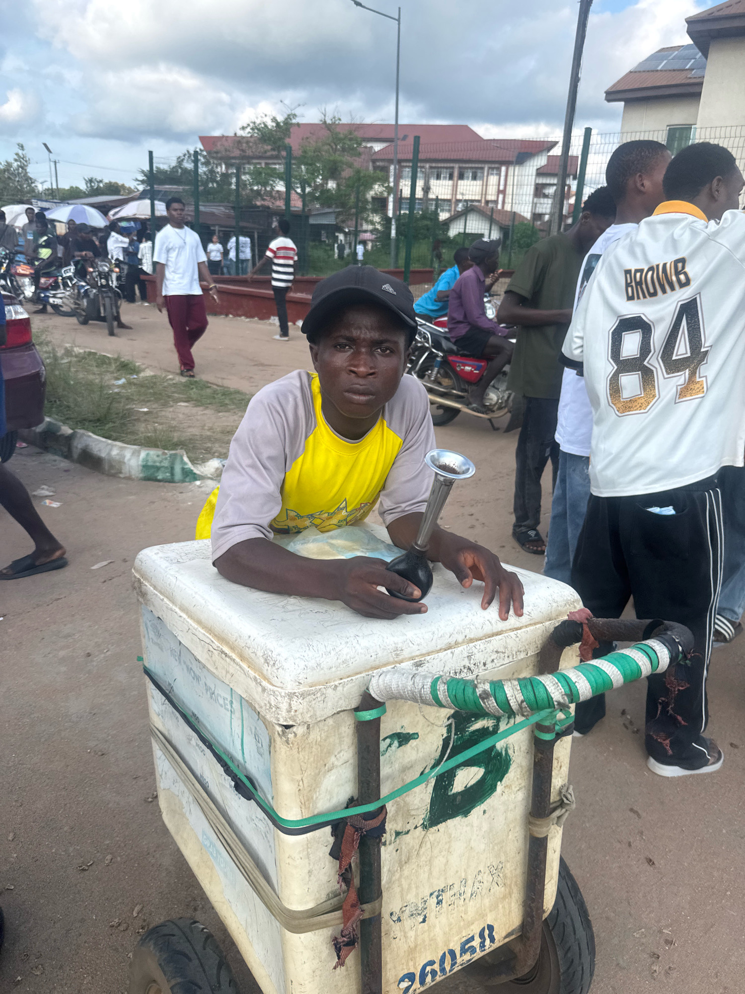 In Ihiagwa, Nigeria, on a cloudy day, a young Black man wearing a gray and yellow t-shirt and a black baseball cap is positioned behind a white cooler cart. The cart, which appears to be used for selling goods, has various straps and markings, including the number "26058" visible on its side. The man is holding a horn-like object to his mouth. The background reveals a street scene with several other individuals, motorcycles, and buildings, suggesting a commercial or public area. The ground is unpaved, and there are indications of dried vegetation and debris.