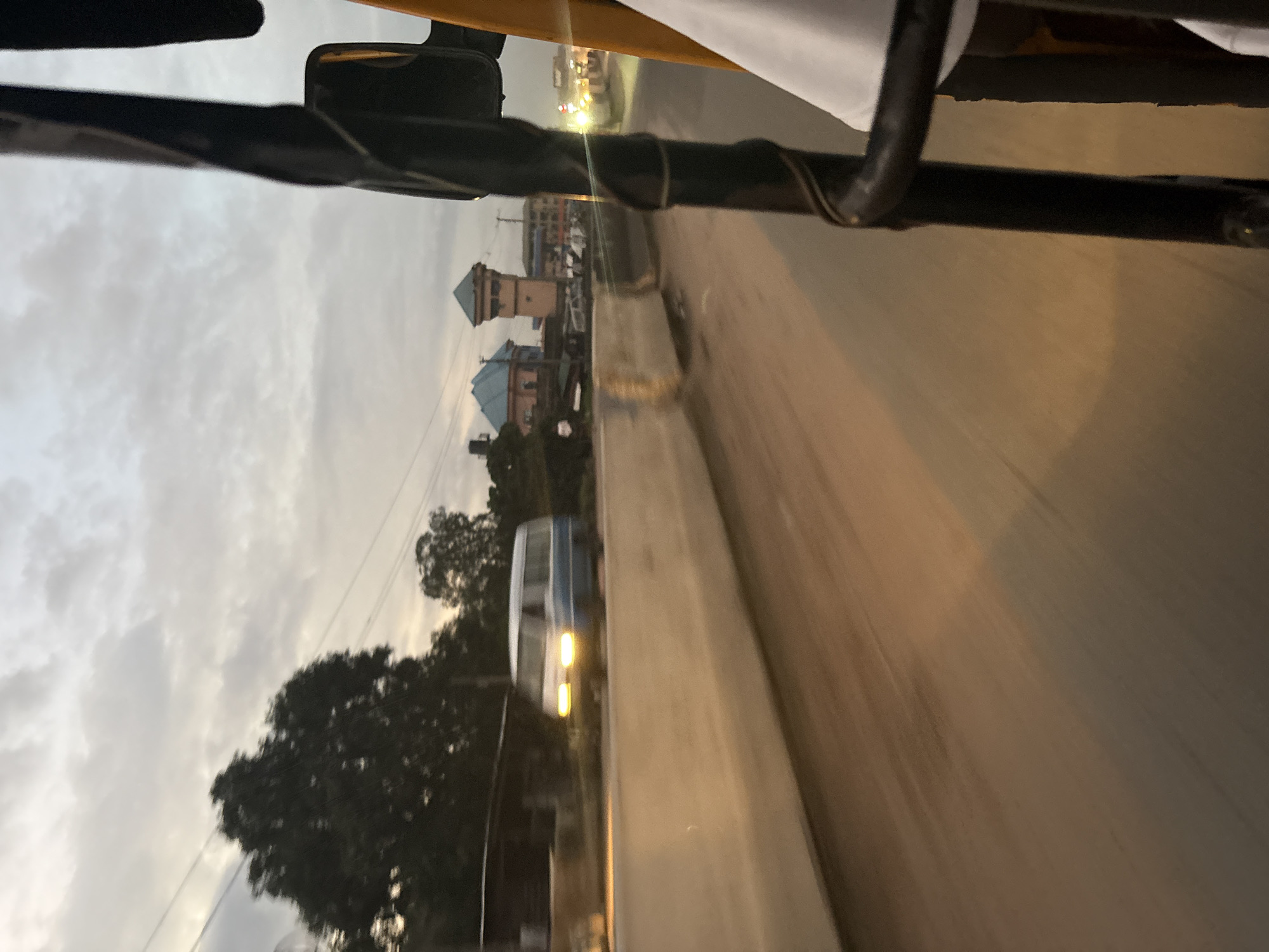 The image depicts a view from the passenger seat of a three-wheeled auto-rickshaw, or "keke," traveling on a road in Onitsha, Nigeria. The sky is overcast with clouds. On the left side of the road, a blue and white minivan with its headlights on is passing by in the opposite direction. In the background, there are buildings, including one with a distinctive tower, and trees. The road surface appears to be paved, and there is a concrete barrier separating the lanes of traffic. The overall lighting suggests it is either early morning or late evening.