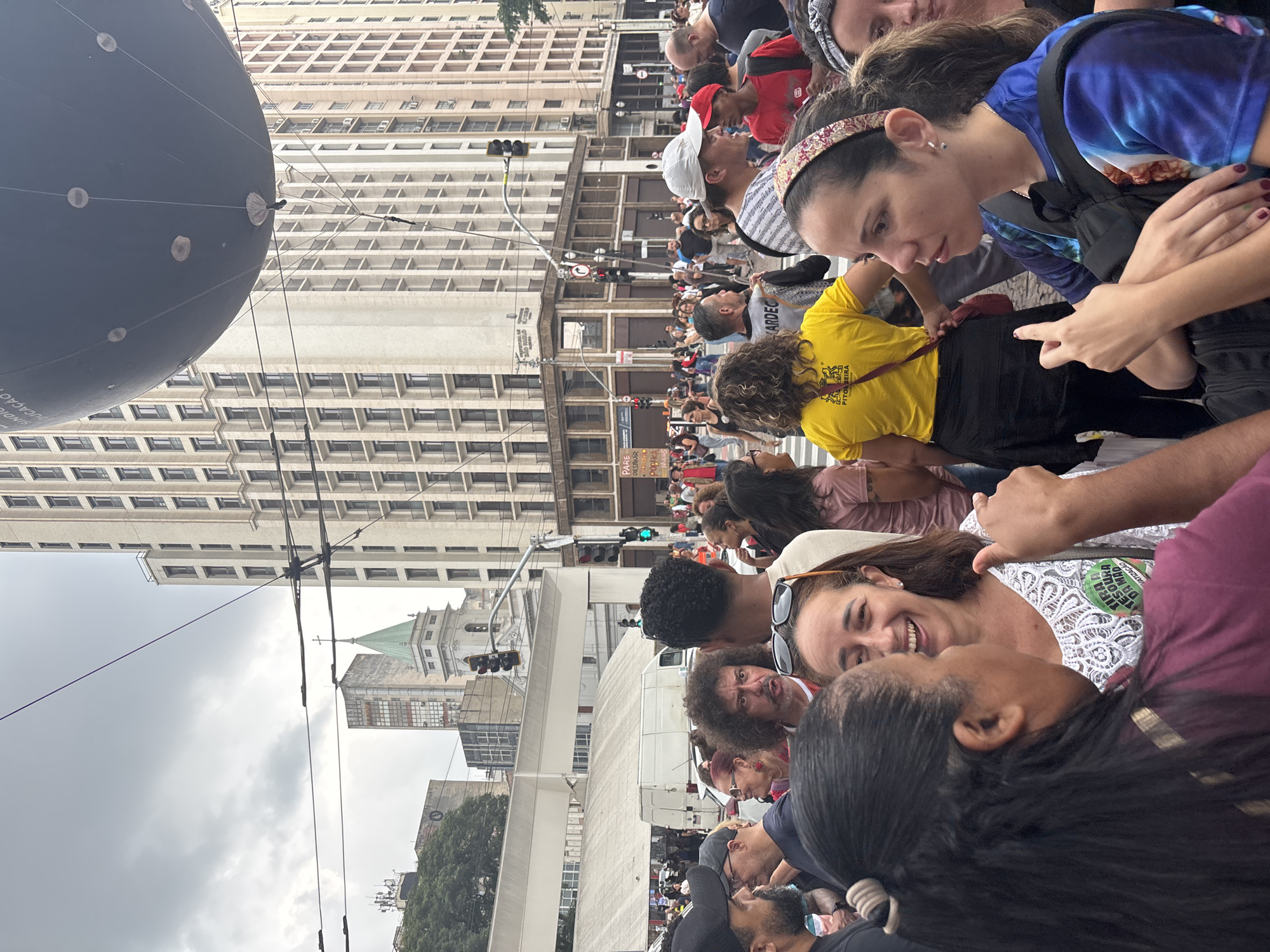 This image captures a vibrant public gathering on a city street in São Paulo, Brazil, under an overcast daytime sky. A large crowd of diverse individuals fills the street, suggesting a demonstration or public event.
In the foreground, a woman wearing a white lace top smiles, adorned with a green badge that partially reads "LIBERDADE DE..." and "MATIZAÇÃO," likely referring to "freedom" and "democratization" or a similar social message. Her gaze is directed towards another individual seen from behind. Nearby, a woman in a blue and white patterned shirt with a dark backpack looks downwards. A man with curly dark hair and a beard is visible in the mid-ground, looking to the right with a focused expression. Another person wears a yellow T-shirt with "PITO BEIRA" printed on the back.
Dominating the left side of the frame is a large, dark gray spherical balloon, tethered above the street. Partially visible text on its surface reads "SINDICATOS," indicating an association with labor unions. The background features a dense urban landscape of tall, multi-story buildings with numerous windows. In the distance, a distinctive building with a green-domed spire, possibly a church, stands out. Multiple traffic lights and a network of overhead power lines or trolley lines crisscross the streetscape. A faint sign in the background partially reads "PARE DE...", suggesting a protest message. The scene conveys a lively, engaged public assembly within a bustling metropolitan environment.