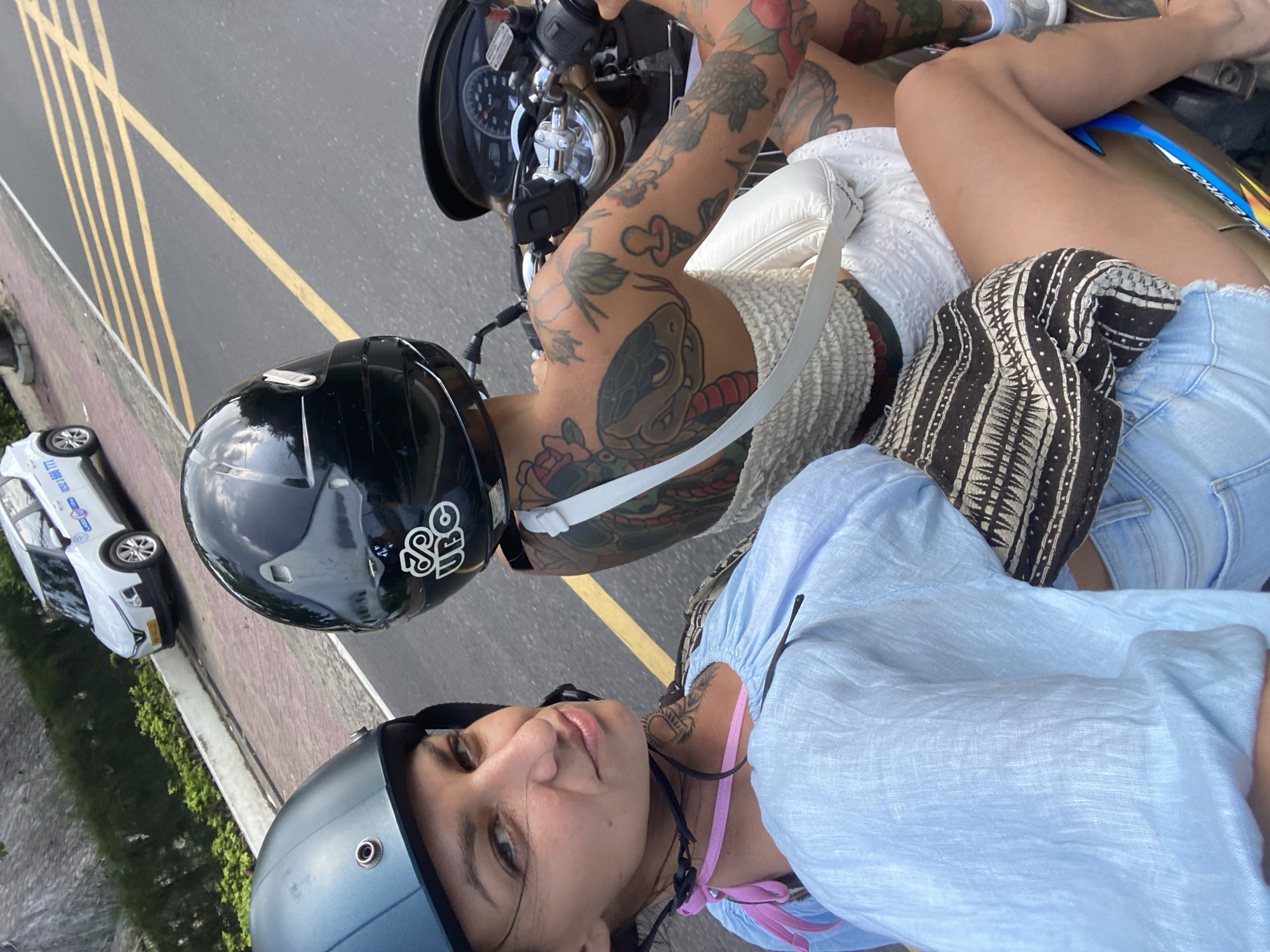 In a city street in Phu Thuy Ward, Vietnam, two individuals are riding on a motorcycle. The person in the foreground, a woman wearing a light blue linen shirt and denim shorts, has a light blue helmet and is looking towards the camera. She has a tattoo on her neck and a pink strap from her garment visible. Behind her, the second individual, wearing a black helmet with a logo, has extensive tattoos covering their arms and back, including a snake and a rose. This person is wearing a white, textured top and is operating the motorcycle. A white car is visible on the side of the road behind them, and the road has yellow lane markings. The environment appears to be a sunny day.