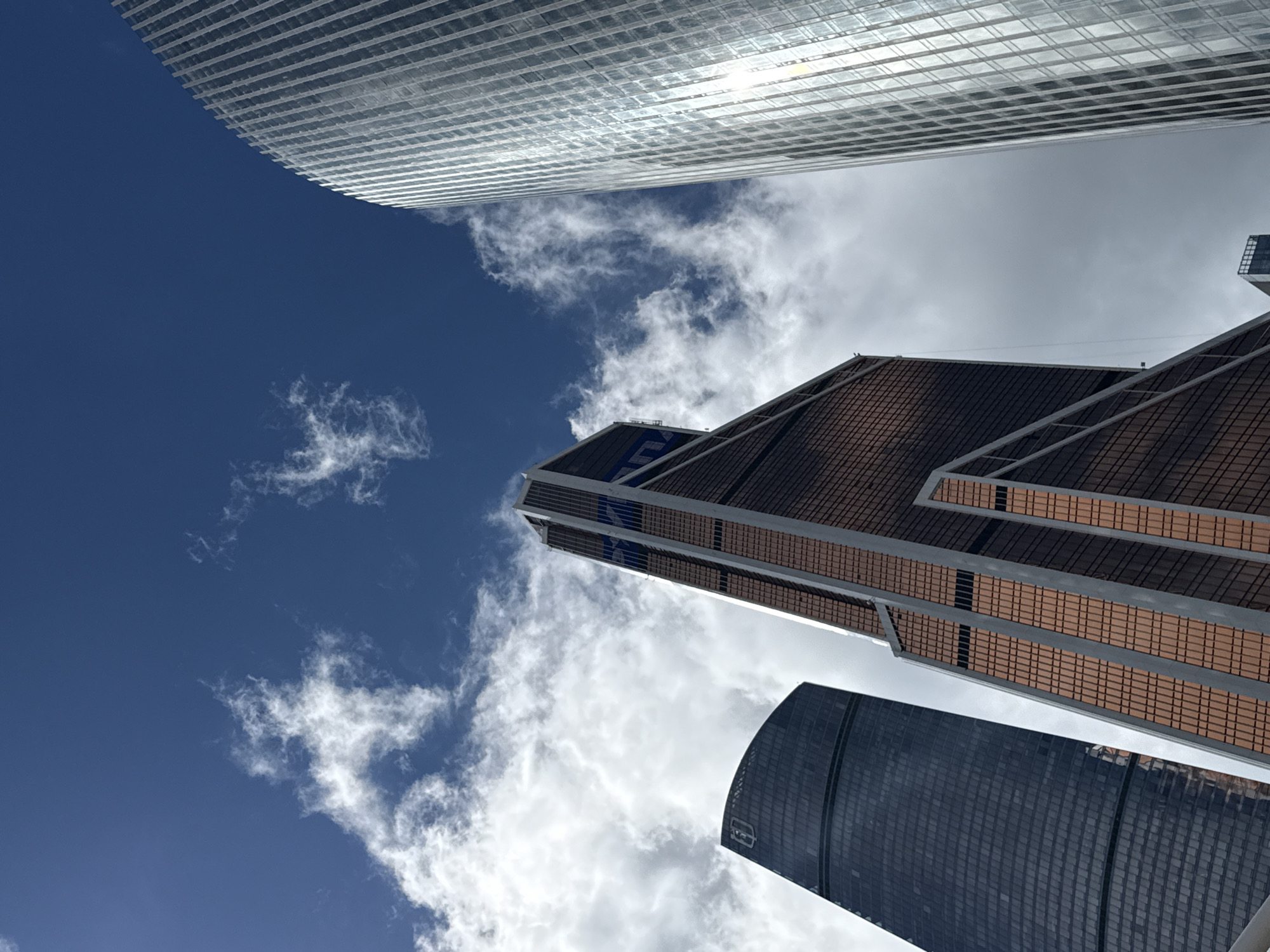 The image depicts a low-angle view of several modern skyscrapers against a bright blue sky with scattered white clouds. The buildings are tall and geometric, with various architectural styles. One prominent building in the center has a reddish-brown facade with vertical dark accents and a pointed top. To its left, a dark, cylindrical skyscraper with a reflective surface is visible. On the right, a very tall, slender skyscraper with a shimmering, silver-like facade made of many vertical panels dominates the frame. The buildings are likely part of a business district, given their scale and contemporary design. The image appears to be taken on a clear day, with sunlight glinting off the glass surfaces of the buildings. The location is likely the Moscow International Business Center (MIBC), also known as Moscow City, in Moscow, Russia, a prominent hub for business and residential skyscrapers. No people are visible in the image.