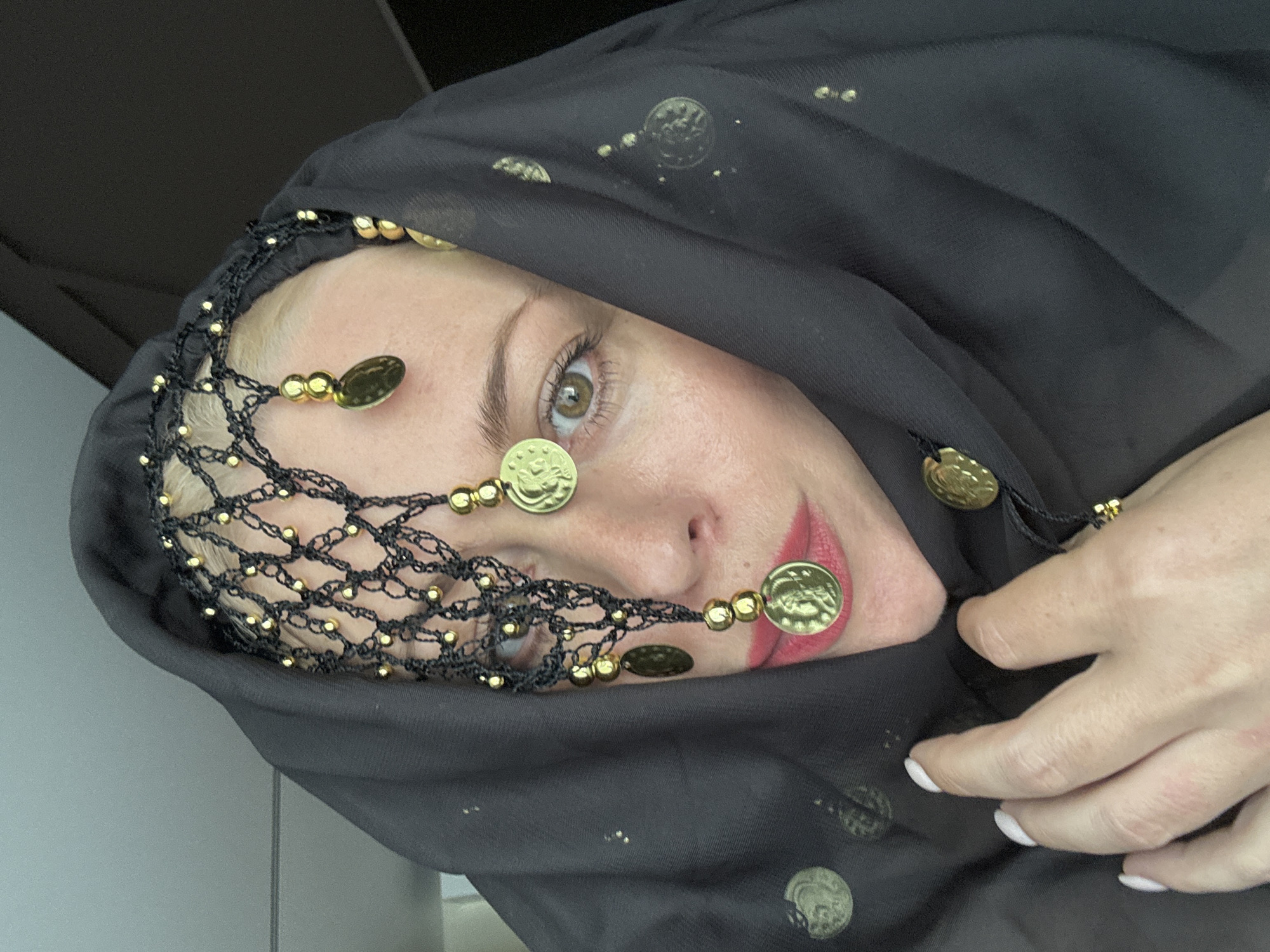 This media file features a close-up, tilted shot of a woman wearing a black headscarf adorned with gold coins and beadwork. The woman has fair skin, light brown eyes, and is wearing red lipstick. The headscarf is partially covering her face, with the intricate beadwork and coins framing her forehead and extending down towards her cheeks. Some of the gold coins are also visible on the fabric of the headscarf itself. A hand with white-painted fingernails is resting on the headscarf, partially obscuring the lower part of the image. The background is dark and out of focus, offering no specific environmental details. The lighting suggests an indoor setting, possibly illuminated by artificial light, with no clear indication of time of day or weather. The scene appears to be a portrait with a focus on the woman's adornments and her gaze. There is no visible text in the image. The provided location context is Sochi, Russia, but this cannot be visually confirmed from the image alone.