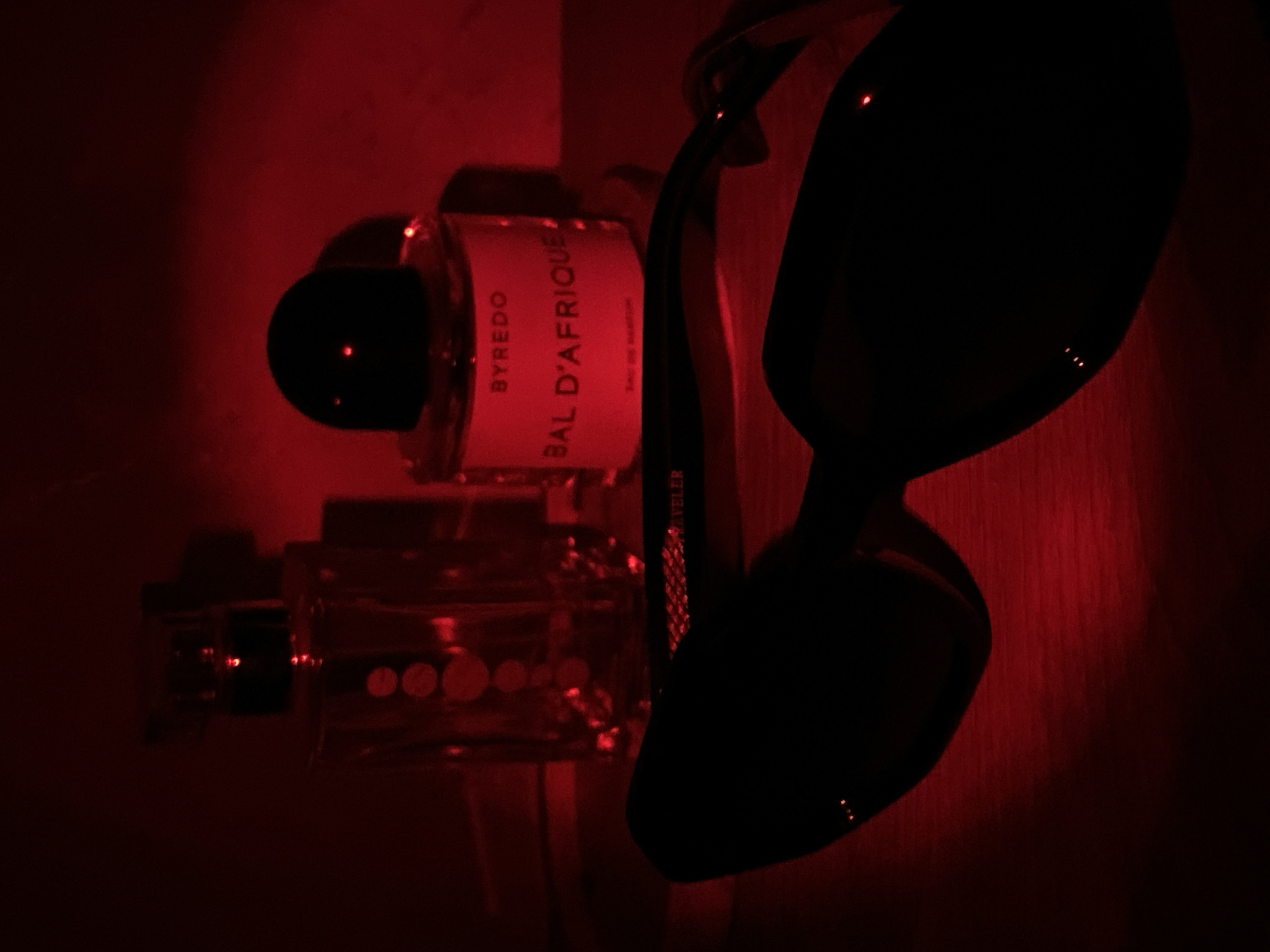This media file presents an indoor still life, bathed in a rich, dark red light, suggestive of an evening or nighttime setting in Sochi, Russia. The background appears to be a wall or textured surface, rendered in shades of deep red.
The primary subjects include two perfume bottles and a pair of sunglasses. On the left, a prominent perfume bottle with a black cylindrical cap is clearly visible. Its label, illuminated by the red light, distinctly displays "BYREDO" at the top, "BAL D'AFRIQUE" in large capital letters below, and "EAU DE PARFUM" at the bottom. The glass of this bottle subtly reflects the red ambient light.
Below and slightly behind the Byredo bottle, another perfume bottle is partially visible. This bottle is squarer in shape, has a dark cap, and features a pattern of small, circular elements on its front surface that catch the red light.
To the right, a pair of dark sunglasses is positioned, largely silhouetted against the red background. The lenses are black and exhibit several small, bright reflections, indicating a glossy surface. Faint, barely legible text is visible on the arm of the sunglasses, which appears to read "REVELER," possibly indicating a brand or model.
No activity or human presence is depicted. The scene is characterized by its dramatic red lighting, which creates deep shadows and highlights the reflective qualities of the objects. The composition emphasizes the aesthetic arrangement and textures of these personal items.