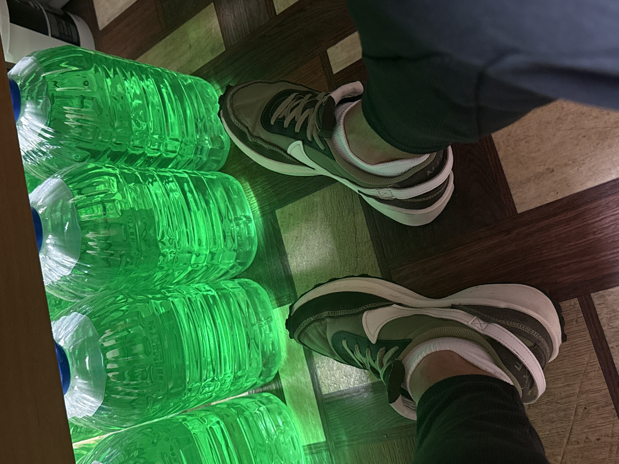 The image shows a person standing on a parquet floor, next to a stack of green-tinted water bottles in a box. The person is wearing grey pants and a pair of beige, green and white Nike shoes with white socks visible. The floor has a pattern of alternating light and dark wood squares. The green tint on the water bottles suggests artificial lighting. The location in the city of Chaikovsky, Russia is not evident in this image.