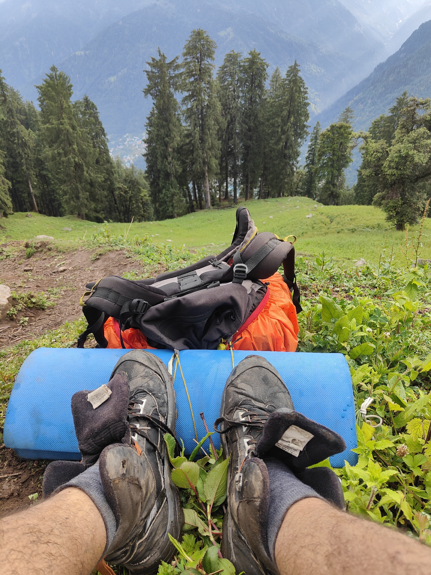 In Vashisht, India, a close-up perspective shows a pair of worn, dark hiking boots on a blue sleeping mat. The boots are partially covered by gray socks, and one foot is positioned over the other. Behind the boots, a black and orange backpack rests on the ground, suggesting a break during an outdoor excursion. The surrounding environment features lush green vegetation and a dirt path. In the background, a dense forest of tall pine trees climbs up a misty mountainside, with a hazy valley visible further in the distance. The overall scene conveys a sense of rest amidst a natural, mountainous landscape.