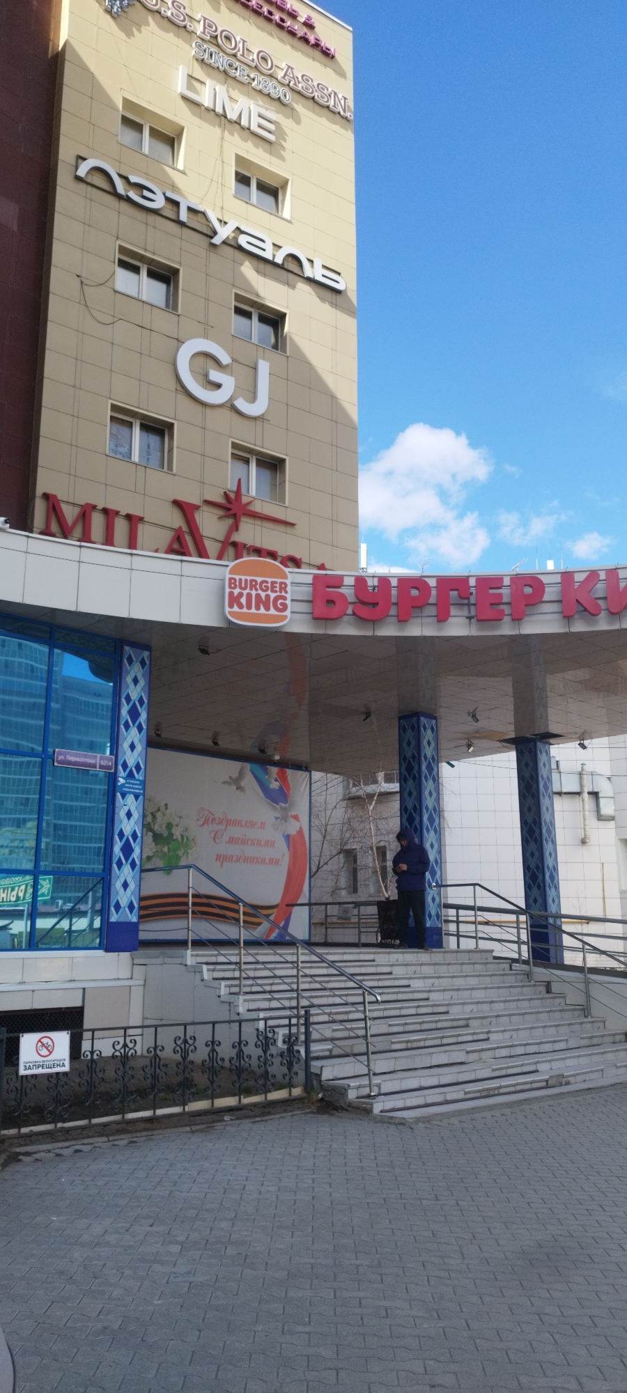This is a photograph of the Burger King restaurant in the city of Yakutsk, Russia. The Burger King sign, written in both English and Russian (БУРГЕР КИ), is visible above the entrance. To the left is a sign for "Milavitsa". The building also has signs for "Lime", "Л'Этуаль" and "GJ". A poster with a patriotic message related to May Day (a holiday often associated with spring and labor) is prominently displayed on the entrance wall. A man in a blue hoodie stands on the steps leading to the entrance. A sign prohibiting bicycle riding is at the bottom of the stairs. The sky is blue with a few clouds. The architecture appears to be a mix of modern and older styles.