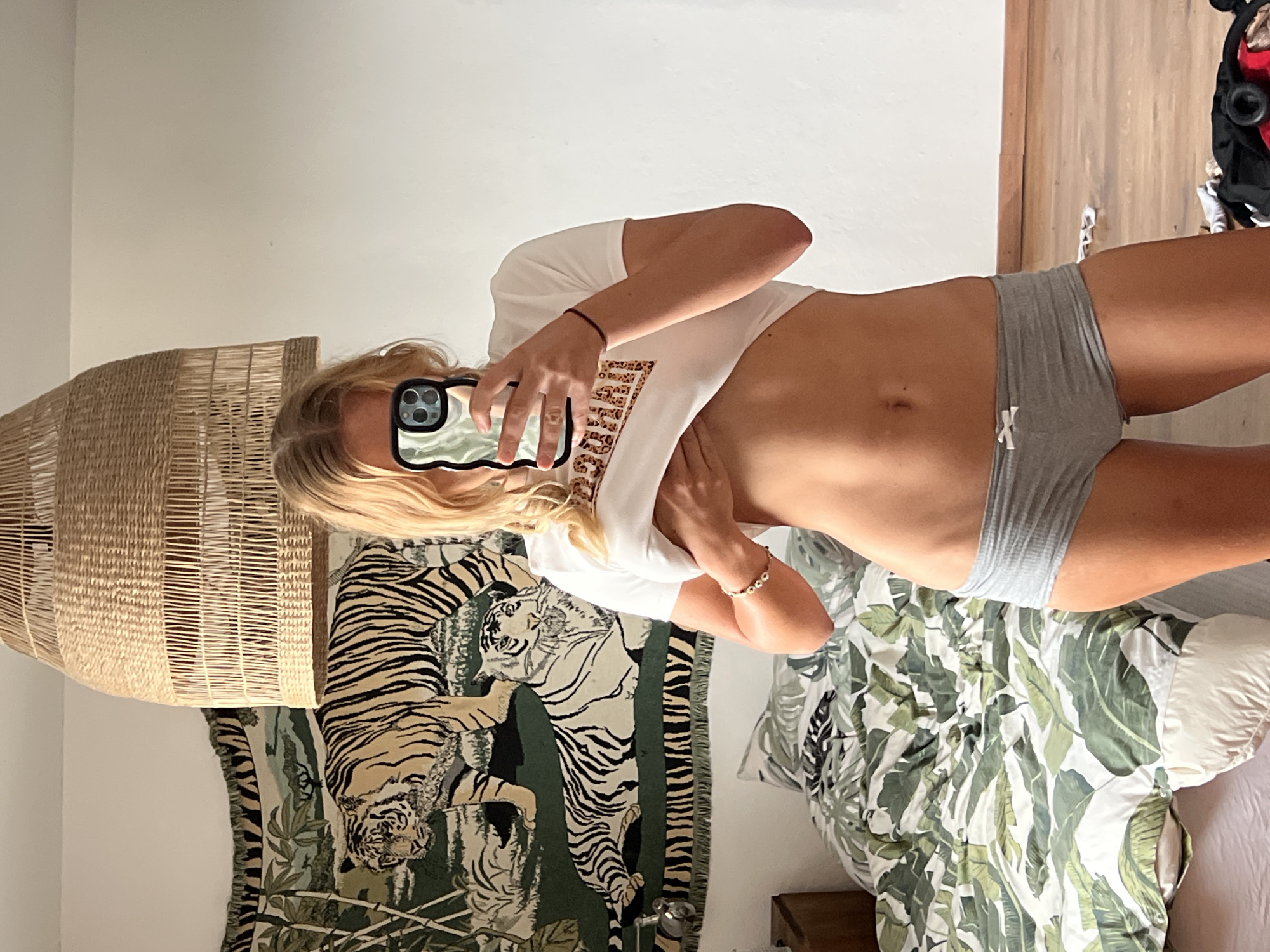 The image shows a woman taking a selfie in a bedroom. She is wearing a white t-shirt and grey underwear. She has blonde hair and is holding a phone in her hand, which is reflecting the surroundings. The bedroom appears to be decorated with a tapestry of tigers and a woven lampshade. There is a bed with a tropical-patterned duvet. The lighting suggests it is daytime. The overall mood is casual and relaxed. No text is visible in the image. The location context of Mühldorf a. Inn, Germany is not evident from the image itself.