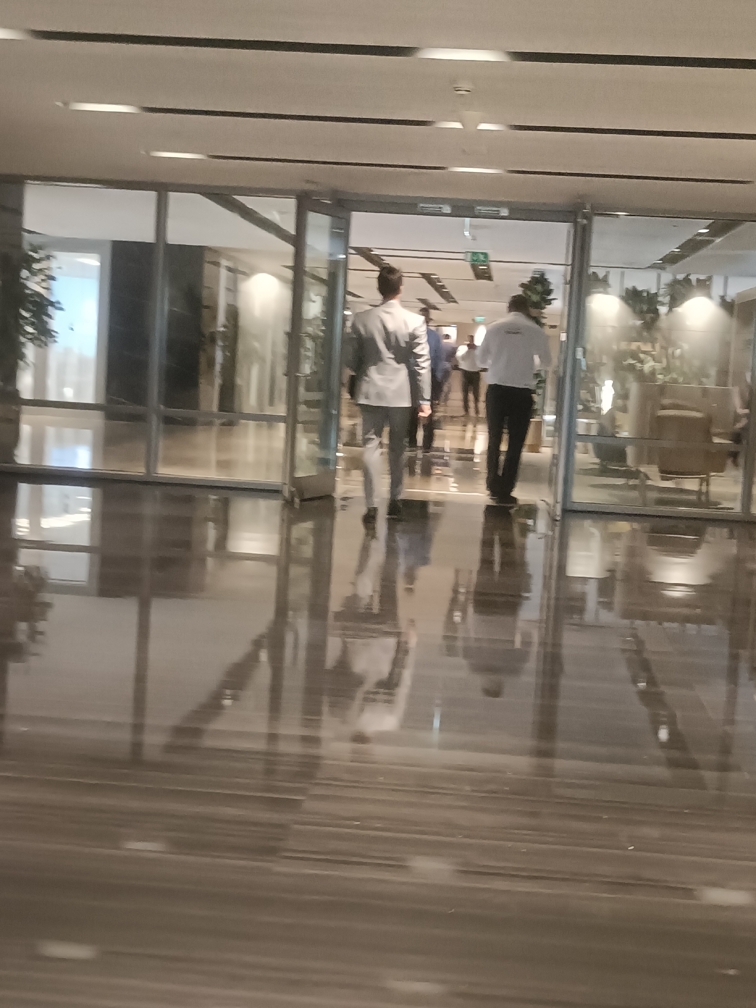 The image shows the interior of a modern building, likely a hotel or corporate office, in Dubai, United Arab Emirates. Two men are walking away from the camera towards an open doorway. The man in the foreground is wearing a light-colored suit and dark pants, while the man behind him is wearing a white shirt and dark pants. The floor is highly polished and reflects the ceiling lights and the people. Large glass windows line the walls, revealing the interior spaces of other rooms. The overall impression is one of a clean, contemporary, and well-lit environment.