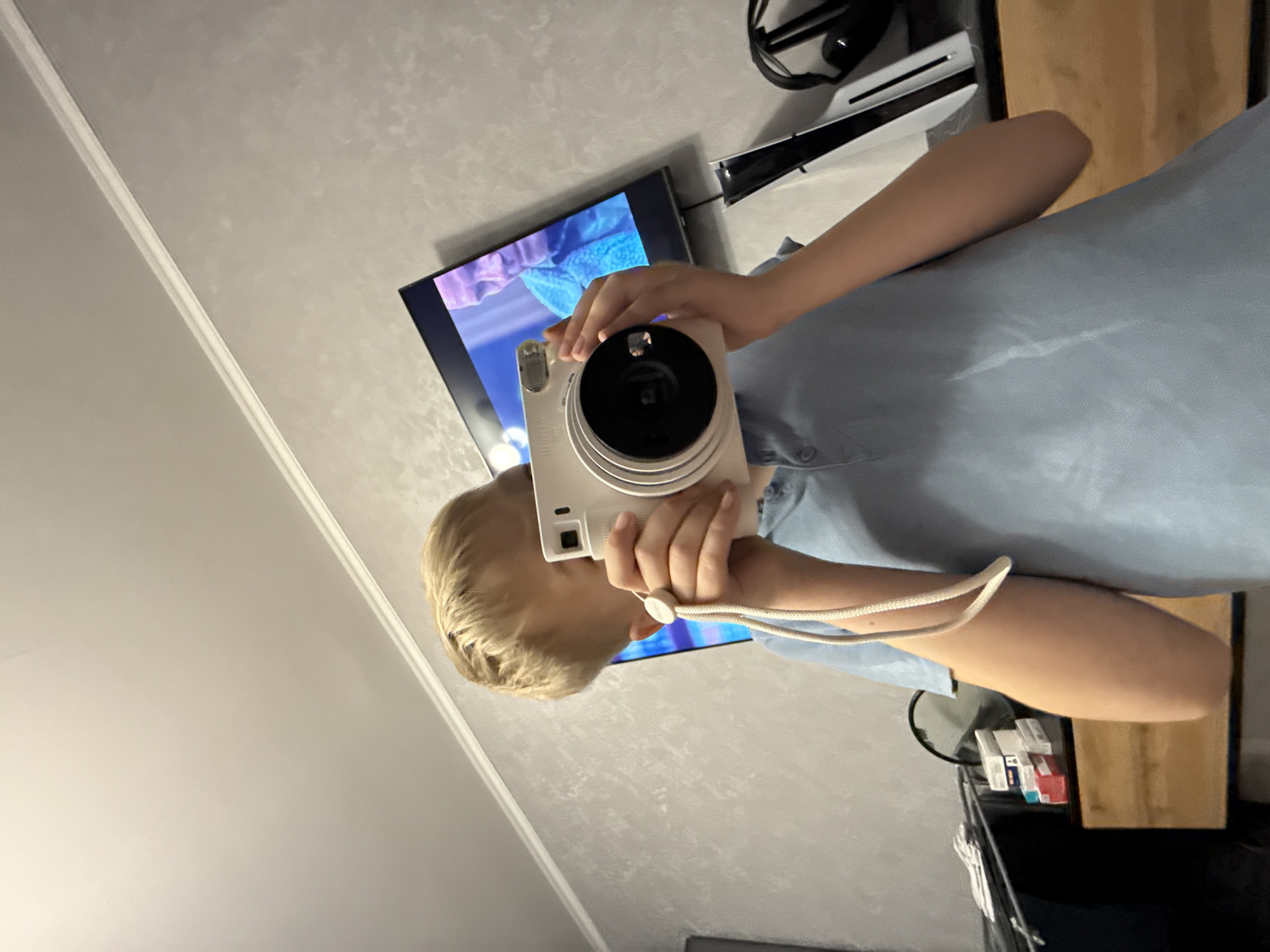 A person with blonde hair is holding a white instant camera in front of their face, obscuring it from view. They are wearing a light blue shirt. Behind them, there is a television displaying a colorful image with blue and purple tones. A white PlayStation 5 console and black headphones are visible on a surface next to the TV. The background suggests an indoor environment with patterned wallpaper. Located in the city of Yakhroma, Russia.