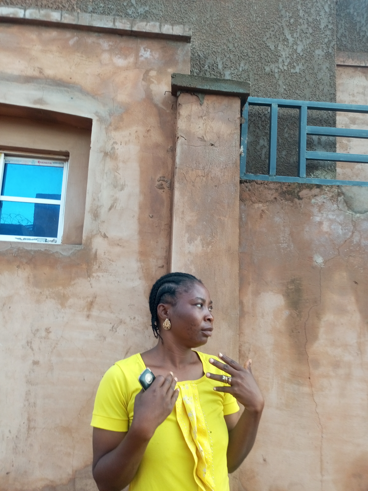 A Black woman is captured in a medium shot, standing in front of a rough-textured, pale brown wall, possibly in Tempure, Nigeria. She is wearing a vibrant yellow short-sleeved t-shirt with a decorative, ruffled placket that features small embroidered dots. Her left hand, adorned with rings, is raised near her chest, fingers splayed as if gesturing or in mid-movement. She holds a dark rectangular object, likely a mobile phone, in her right hand. Her hair is styled in braids pulled back from her face, and she wears a prominent gold earring in her visible left ear. To her left, a small window with a blue reflective pane is visible, set within the wall. Behind her, a dark blue metal railing separates the wall from a textured grey upper section of the building. The overall setting appears to be an outdoor urban environment.