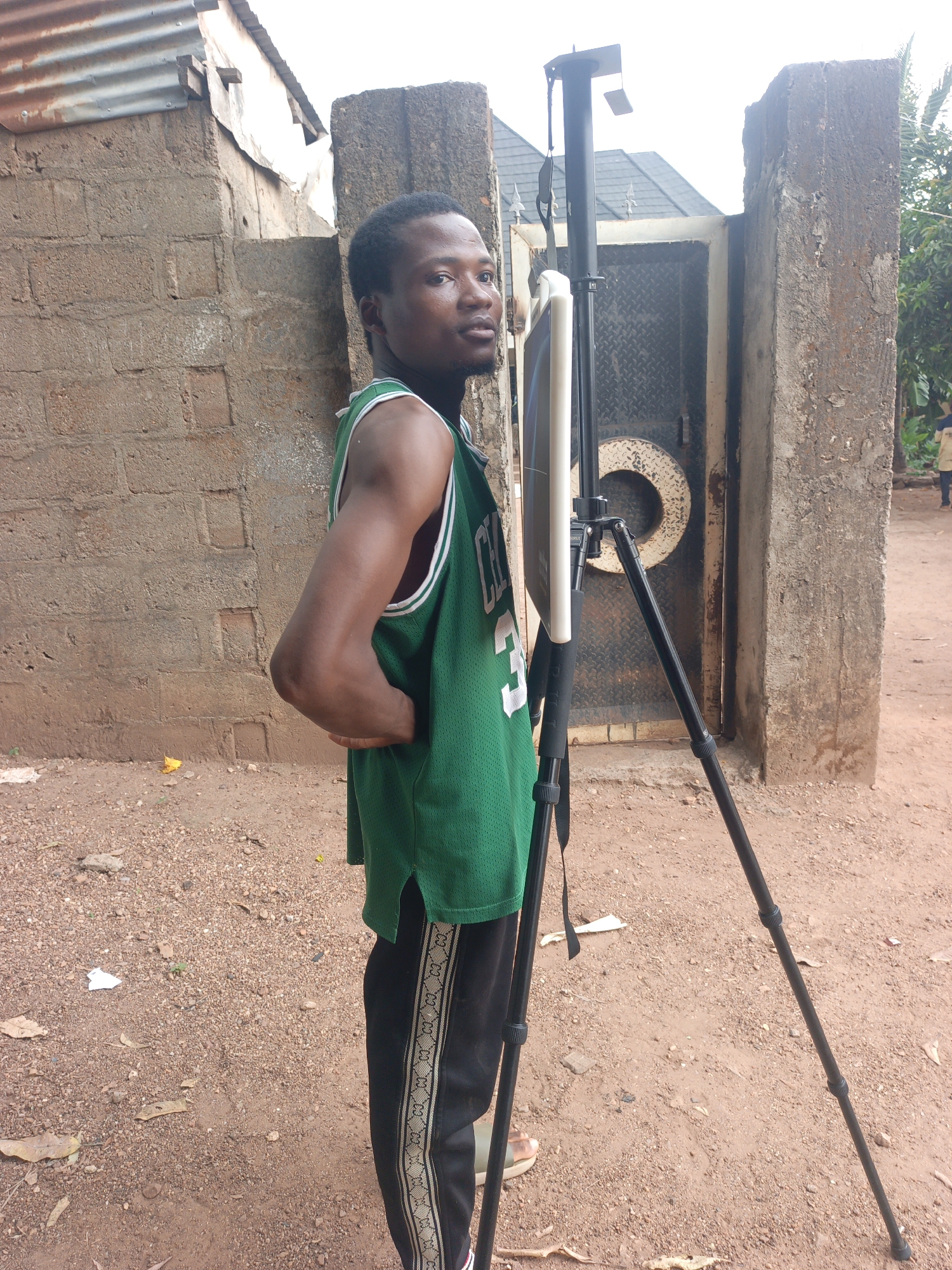 A medium-shot photograph captures a man wearing a green sports jersey, black pants, and sandals. He stands on red dirt amidst gravel and assorted debris next to a tripod holding a white reflector, with a gated concrete wall behind him. The location is Akwanga, Nigeria. A concrete building with a corrugated metal roof is on the left, while foliage can be seen on the right.