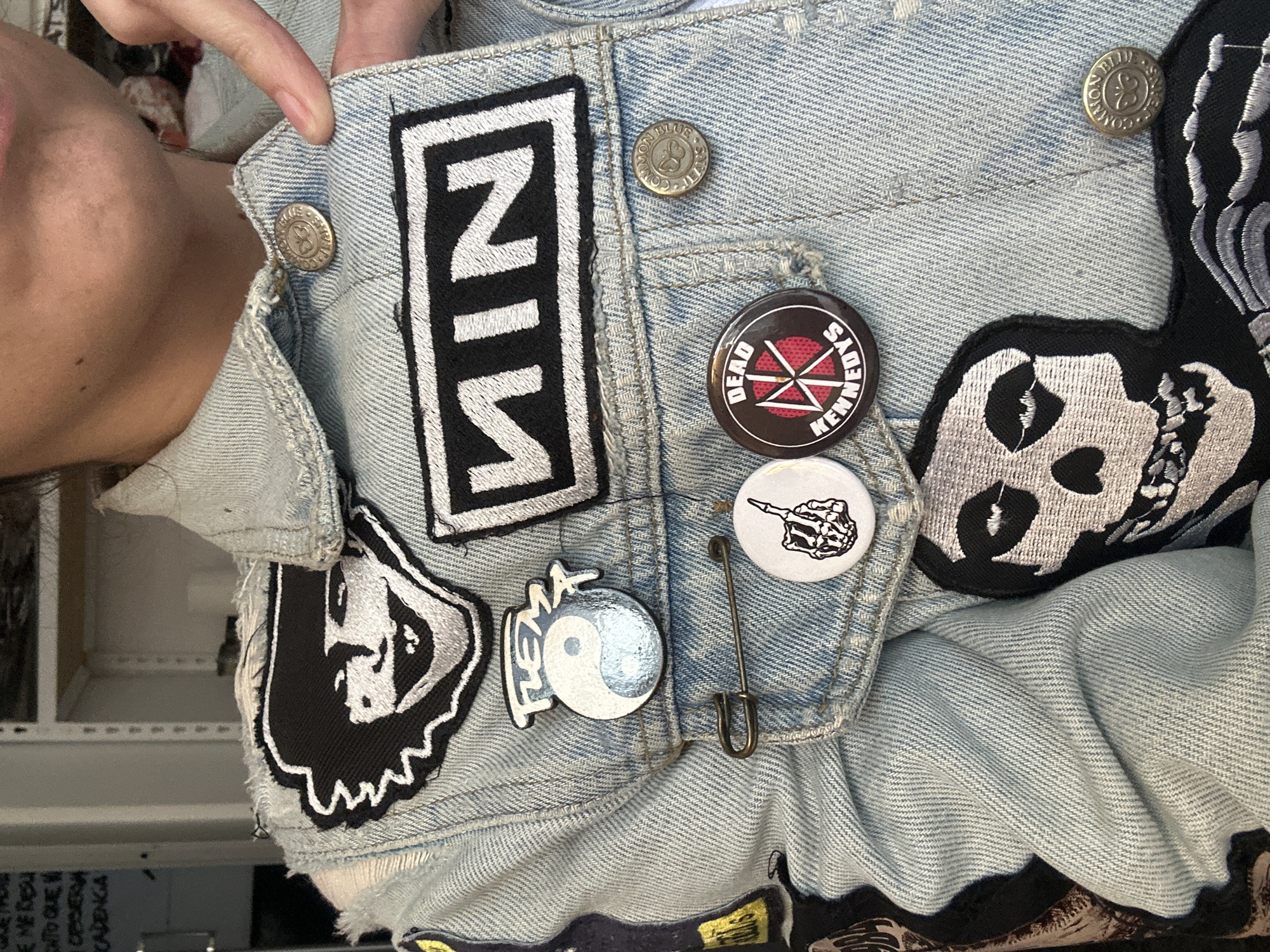 Close-up of a person wearing a light blue denim jacket with various patches and pins. Patches include a "NIN" logo, a skull with a heart-shaped eye, and a stylized portrait. Pins feature a Dead Kennedys logo, a skeleton hand flipping the middle finger, and a yin-yang symbol with the word "FLEMA" written above. A safety pin is also present. The jacket has metal buttons, and part of the person's face is visible. The location is Buenos Aires, Argentina.