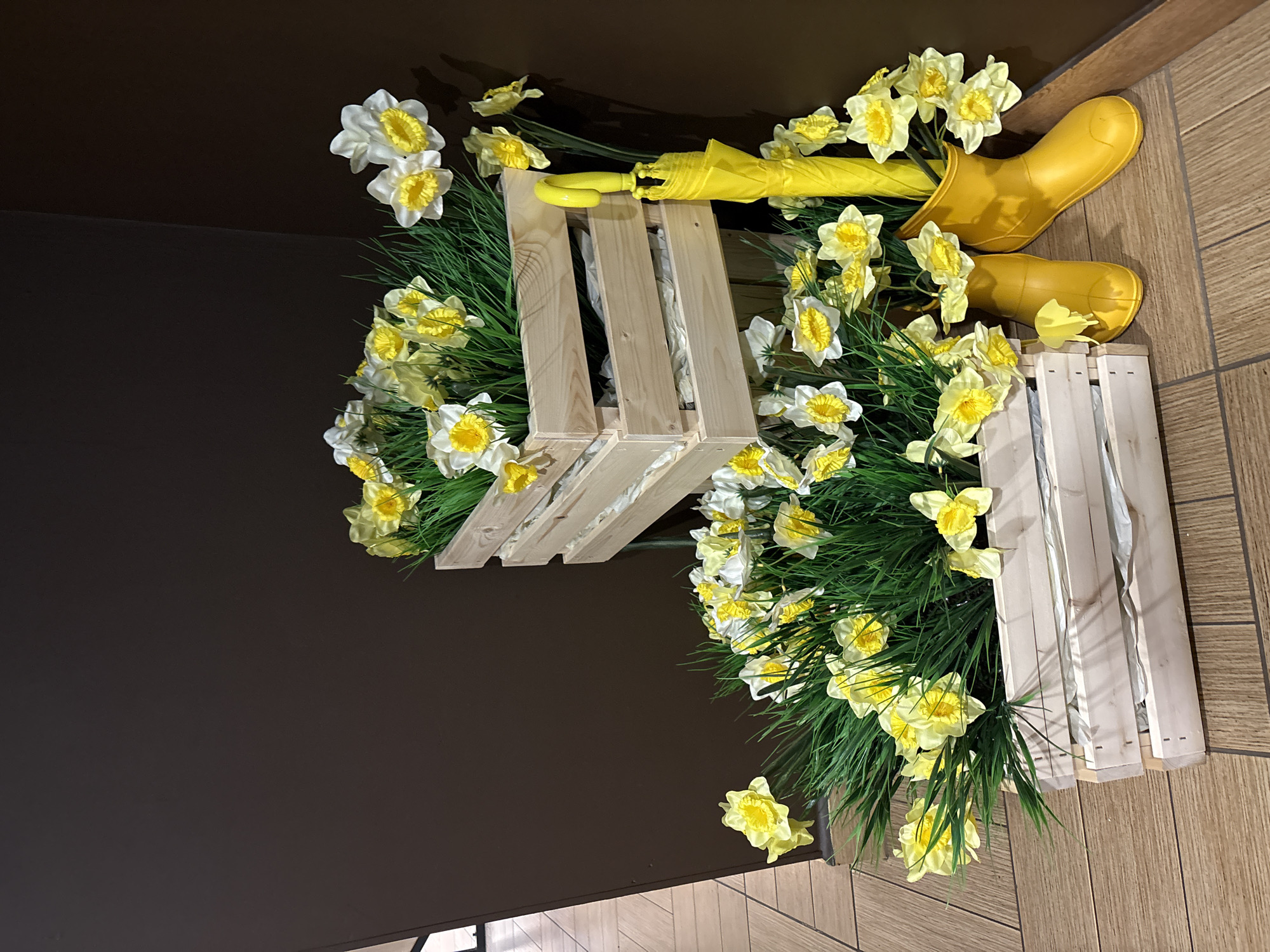 This is a still image of a decorative display featuring faux daffodils and artificial grass arranged in wooden crates. The display is placed next to a dark brown wall on a tiled floor. Two yellow rubber boots and a yellow umbrella are also part of the arrangement. The image does not contain any people, nor does it provide any information about its location in the city of Chaikovsky, Russia, or any specific actions or interactions. The lighting suggests an indoor setting, possibly a shop or a public space.