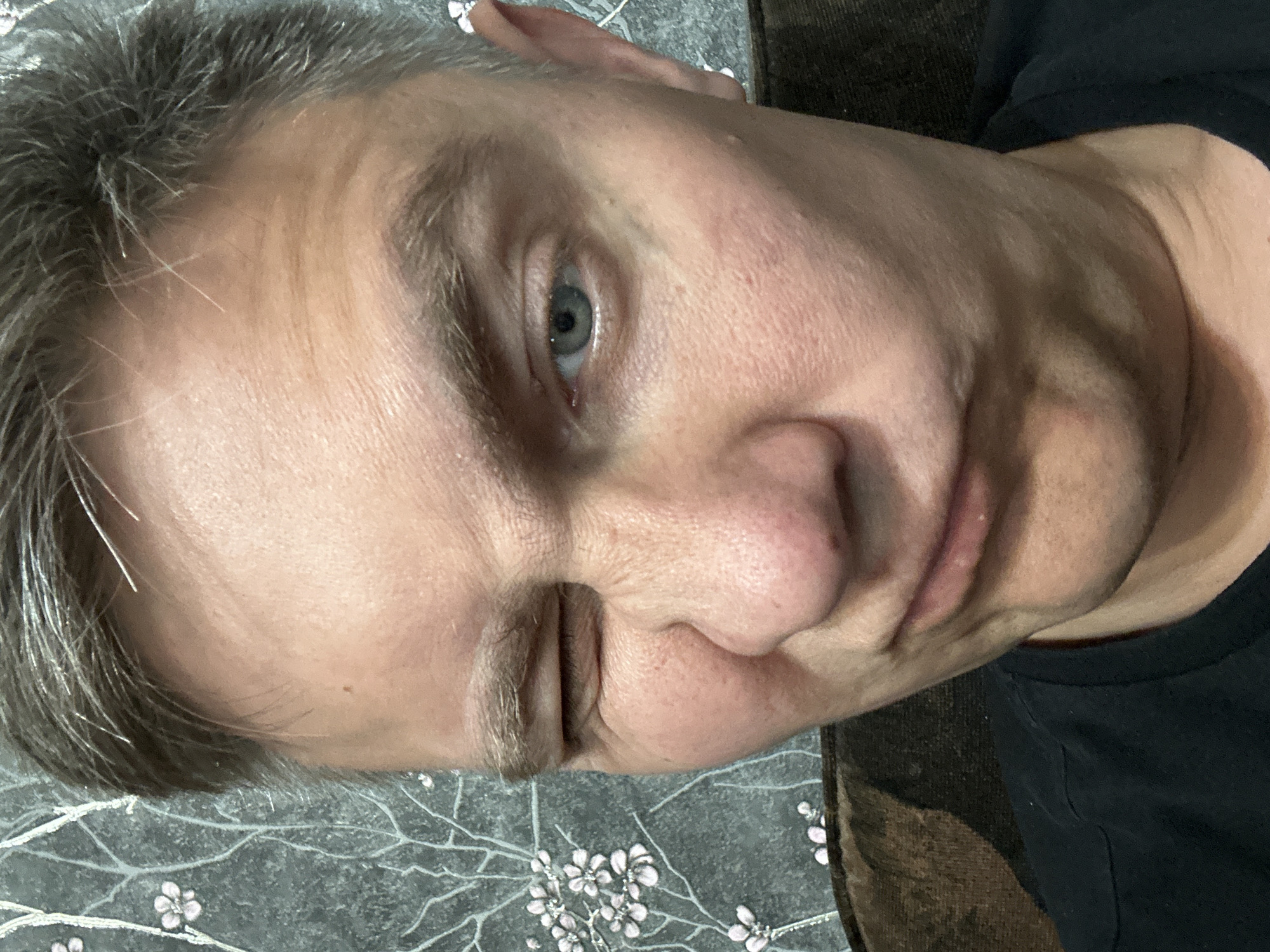 A close-up photograph shows a middle-aged Caucasian male with short, graying hair. His right eye is open and blue, while his left eye is closed or squinted shut. The man has fair skin with visible pores and wrinkles. He is wearing a dark-colored shirt. The background includes a surface with a floral pattern and part of a brown surface. There is no evidence to connect this photo with the city of Minsk, Belarus.