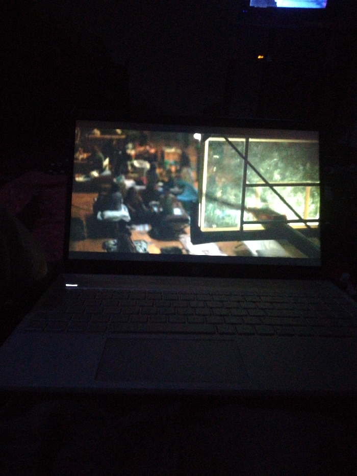 A laptop screen illuminates a dark room. The display shows a scene: a room with people gathered around a table. To the right, a window with a unique, possibly decorative, frame allows a view of a green, leafy outdoor area. The laptop keyboard and trackpad are visible in the foreground, adding to the low light environment. Another television or monitor is visible in the upper center of the image.