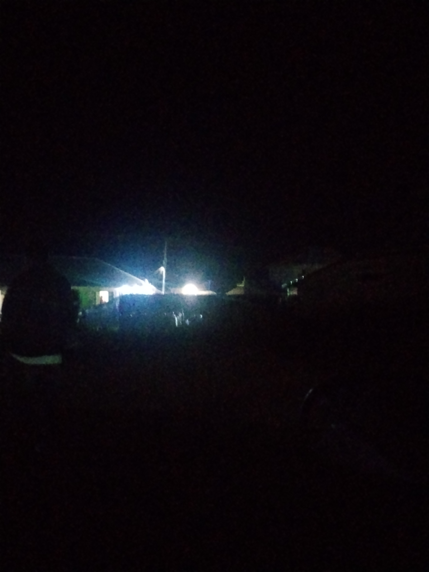 The image depicts a dimly lit outdoor scene at night, possibly in Durumi, Nigeria. The foreground is mostly dark, with the silhouette of a person's head and shoulders visible on the left. In the mid-ground, a building with a lit interior and exterior lights is visible. Several figures, appearing to be people, are present near the building, some standing and others possibly seated. The environment appears to be a cleared area, possibly a yard or open space, with some indistinct vegetation. Bright artificial lights illuminate the building and its surroundings, creating a stark contrast with the surrounding darkness. There are indications of a gathering or event taking place, suggested by the presence of multiple people and lighting.