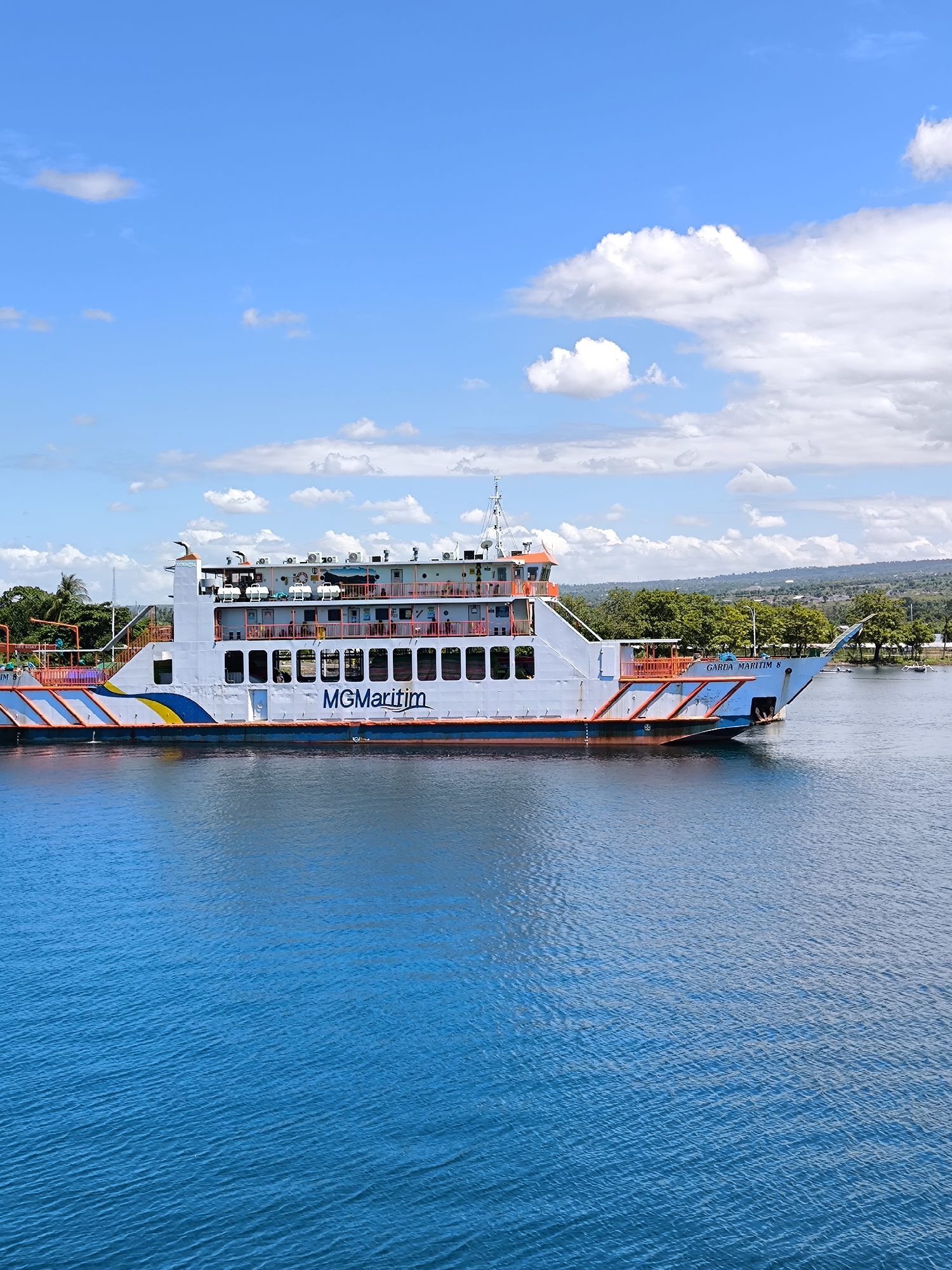 A ferry named "MG Maritim" is sailing on the water in Gunung Malang, Indonesia. The ship is predominantly white with orange and blue accents. It has multiple decks and a prominent antenna. The surrounding water is a vibrant blue. In the background, there is a coastline with green trees. The sky is blue with scattered white clouds.