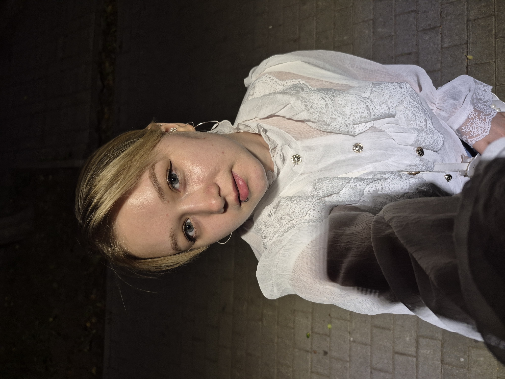 A young woman with blonde hair and blue eyes is shown in a close-up, low-angle shot, with the camera positioned slightly above and to her right. She is wearing a white, sheer blouse with lace detailing on the collar, cuffs, and front. The blouse has a ruffled neckline and decorative buttons. She has a lip ring and hoop earrings. Her gaze is directed towards the camera.
The background consists of a paved walkway with a dark, textured surface, possibly asphalt or concrete, and some faint, blurred greenery to the left. The lighting suggests it is nighttime, with a warm, artificial light source illuminating the woman's face. The scene appears to be outdoors, with the texture of the ground indicating a sidewalk or pedestrian area. No visible text is present in the image. The overall mood is casual and direct, with the subject making eye contact with the viewer. The image is oriented sideways.