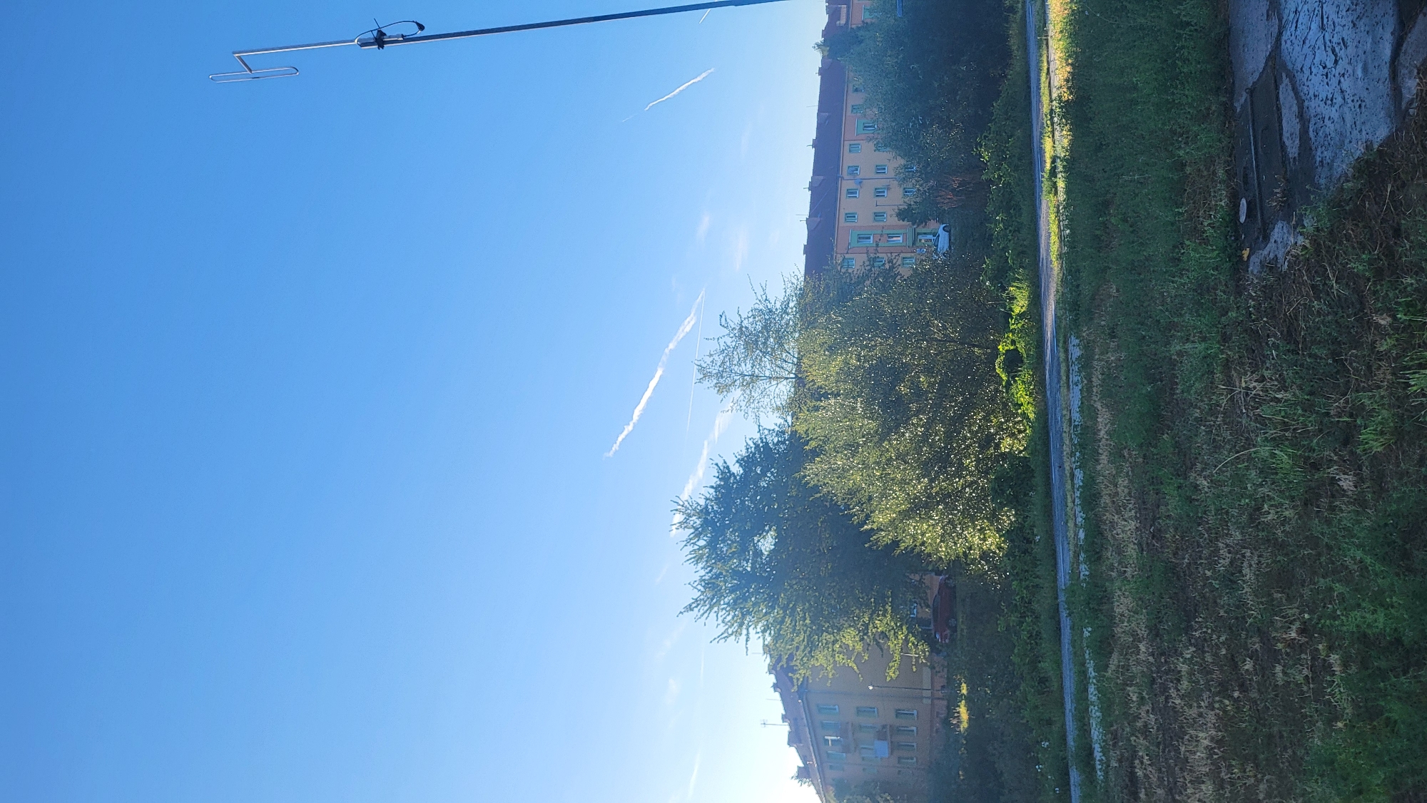 The image depicts a bright, sunny day in Prešov, Slovakia, with a clear blue sky. In the foreground, a patch of rough, green grass and a section of a paved surface are visible. In the middle ground, dense green trees with visible foliage form a backdrop. Behind the trees, a multi-story apartment building with a light-colored facade and orange-tiled roof is situated. A red car is parked on the street in front of the building, partially obscured by trees. A tall antenna or mast with a horizontal element extends from the right side of the image into the sky. Several faint white contrails from airplanes are streaked across the blue sky. The scene appears to be an outdoor urban environment.