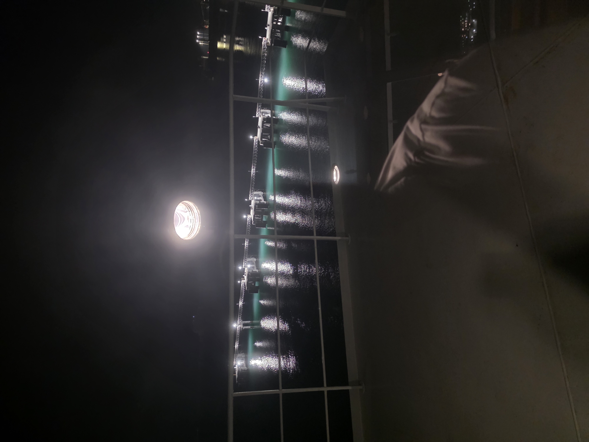 The image shows a night view from a ship's deck towards a pier extending into the water. The pier is illuminated by numerous lights, casting reflections on the turquoise water. The sky is dark, and a bright overhead light fixture is visible in the upper center of the frame. In the foreground, a white railing separates the deck from the view of the pier. A shadowy figure or draped object is present on the deck in the lower right portion of the image. The lights of distant buildings can be seen on the horizon. The scene appears to be captured at night from a vessel docked or moving in the vicinity of Brindisi, Italy, likely near the port area where such piers are common.