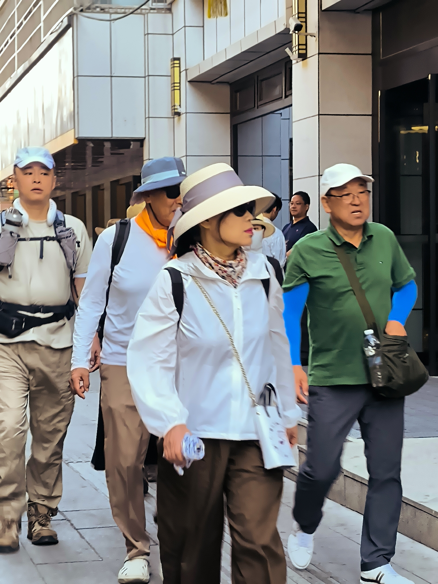 This image captures a street scene in Ürümqi, China, with several individuals walking along a pedestrian walkway. In the foreground, a woman wearing a wide-brimmed hat and a white jacket is walking with a group. She carries a small white purse with a chain strap. To her left, a man in a light blue cap and a beige shirt is also walking, with a backpack and a fanny pack visible. To the right of the woman, a man in a green polo shirt and dark pants is walking, wearing blue arm sleeves and carrying a black bag with a water bottle. Behind him, another man in a white t-shirt and hat can be seen. The background features modern buildings with glass and tiled facades. The walkway appears to be paved with square tiles. The overall impression is of a casual outdoor excursion in an urban setting.