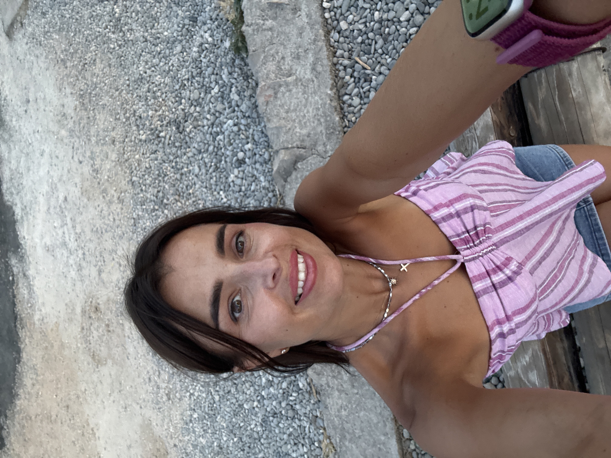 A woman is taking a selfie outdoors. She has dark hair and is smiling, wearing a pink and white striped top and denim shorts. She is also wearing a necklace and a smartwatch. The background appears to be a stony area, possibly a beach or pathway, with a low stone wall. The lighting suggests it is daytime.