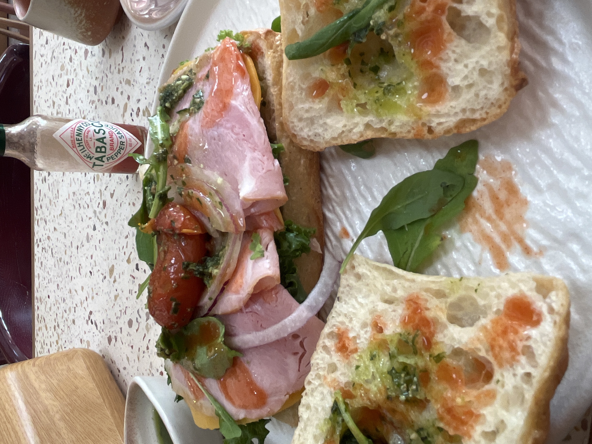 The image shows a partially disassembled sandwich on a white plate. The sandwich appears to be made with ciabatta bread, ham, cheese, tomatoes, red onion, and arugula. A bottle of Tabasco sauce is also visible near the sandwich. The photo appears to be taken on a light-colored, speckled table. There is no evidence that the photograph was taken in Bac Nha Trang Ward, Vietnam.