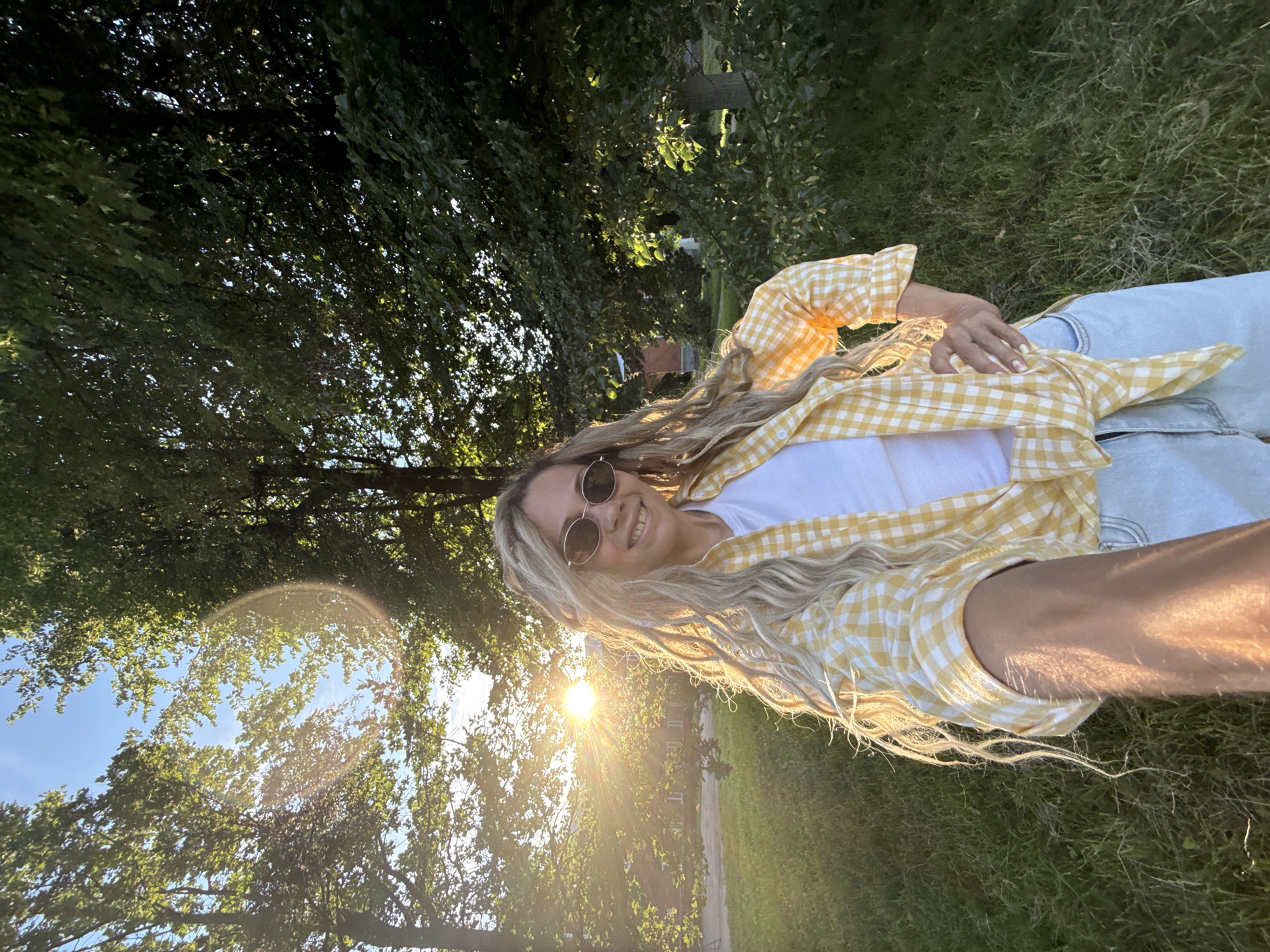 A woman with long, blonde, wavy hair and sunglasses is taking a selfie outdoors in a grassy area with trees. The sun is setting, casting a warm glow on the scene and creating lens flares. She is wearing a white t-shirt, a yellow and white checkered shirt tied at the waist, and light-wash denim shorts. She is smiling and has her left hand on her hip. In the background, there is a building and more trees. The image appears to be taken in a park or a residential area on the outskirts of the city of Шебунино, Russia.
