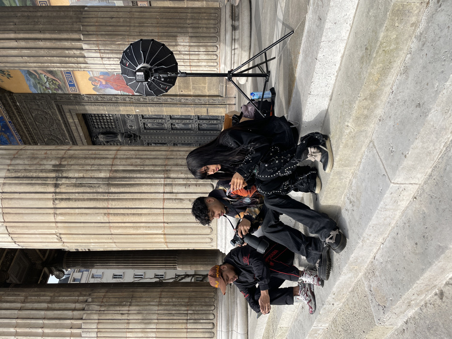Three individuals are seated on the stone steps of L'église de la Madeleine, located in the 8th arrondissement of Paris, France.
From left to right: A male wearing a brown cap, black athletic jacket with red accents, and multi-colored sneakers is looking down at a smartphone held in his left hand. The central individual, a male with dark curly hair and wearing a patterned jacket, is operating a black DSLR camera with a large lens. The rightmost individual, a female with long dark hair and bangs, dressed in a black studded jacket and similarly embellished dark pants, is looking down at a smartphone.
To the right of the group, a black photographic softbox is mounted on a tripod stand. A light blue water bottle and a dark bag are visible on the steps behind the female subject. The background consists of large, fluted classical columns, ornate dark metal doors, and sections of painted murals on the building's stone facade. A multi-story building is partially visible in the far background between the columns. All subjects are independently focused on their electronic devices.