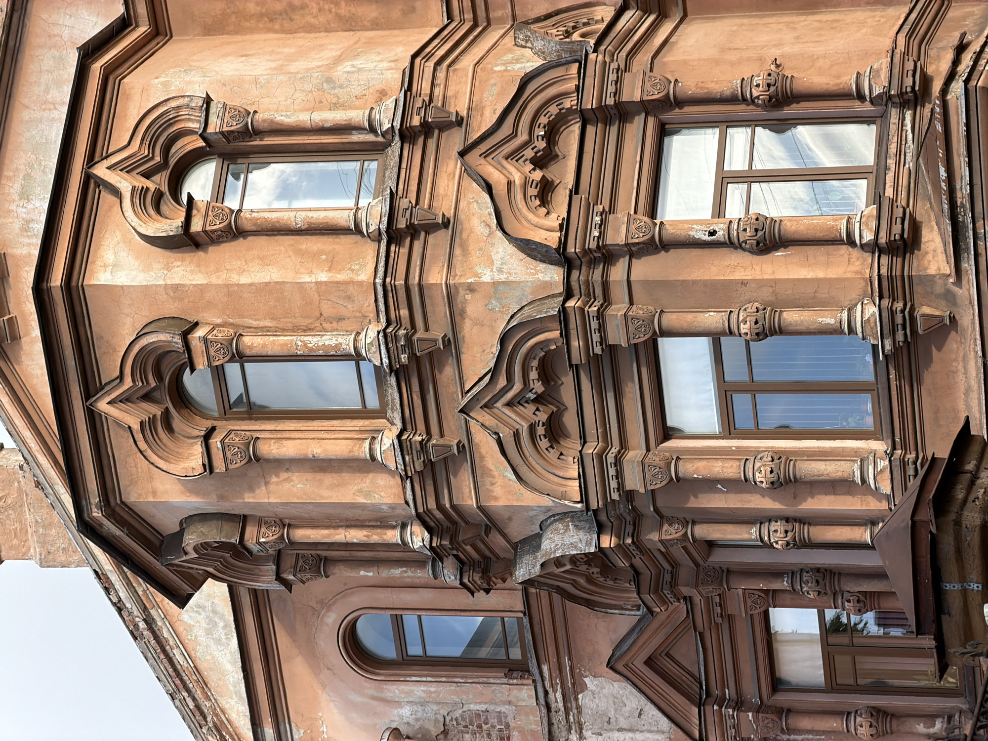 The image shows a portion of a building facade in Saint Petersburg, Russia. The building is characterized by its ornate architectural details. The facade is predominantly an old brick or stone color, with decorative elements around the windows. The windows have an arched or curved design at the top. The architectural details feature intricate designs, including moldings and possibly sculpted elements. Some wear and tear is visible on the facade. The overall impression is one of historical architecture with a blend of grandeur and decay.