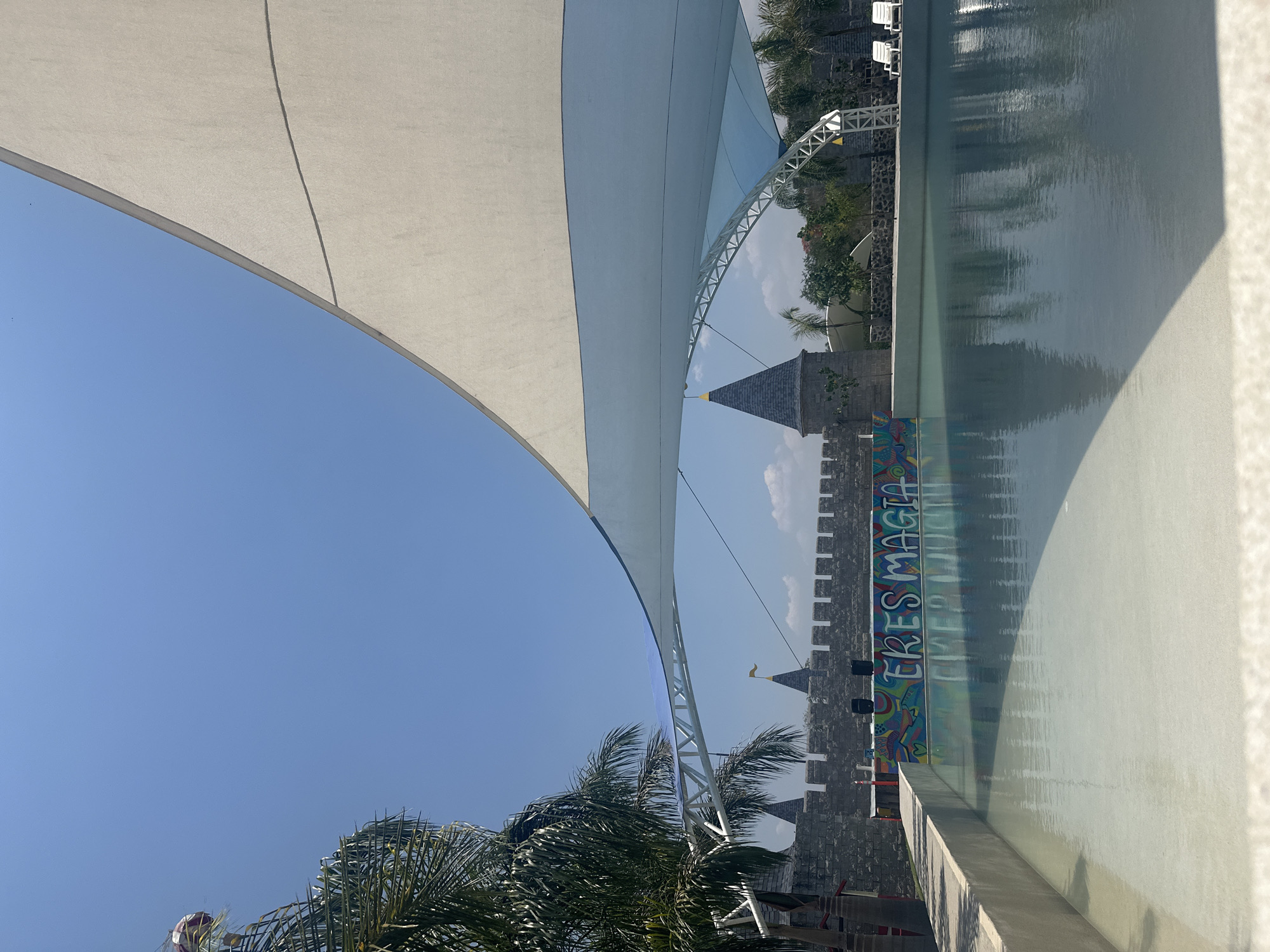 The image shows a castle-themed structure with a mural that says "ERES MAGIA" (You are magic) beside a pool of water. The sky is blue and clear. The presence of palm trees suggests a warm climate. The photograph also shows a structure that appears to be a white fabric sun shade. The image might have been taken in Jicarero, Mexico, based on the user's prompt.