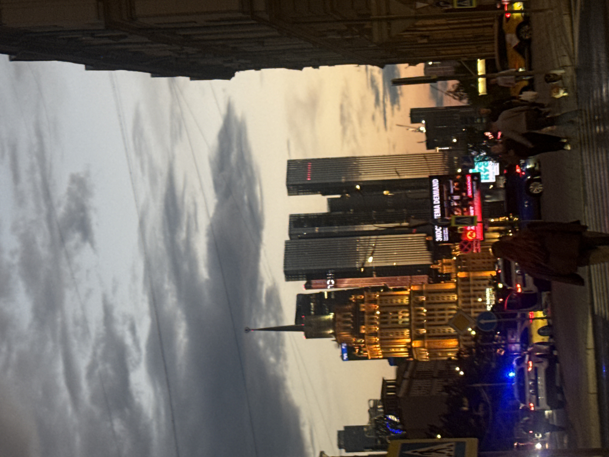 The image depicts a nighttime street scene in Moscow, Russia, with a prominent Stalinist skyscraper in the center, illuminated by warm, yellow lights. The sky is a twilight hue with dark, cloudy formations. In the foreground, a police car with flashing blue lights is visible, suggesting a recent or ongoing incident. Several other vehicles, including a yellow car, are also present on the street. Pedestrians are seen walking, some with their backs to the camera. The overall atmosphere is urban and bustling, typical of a city at night.
