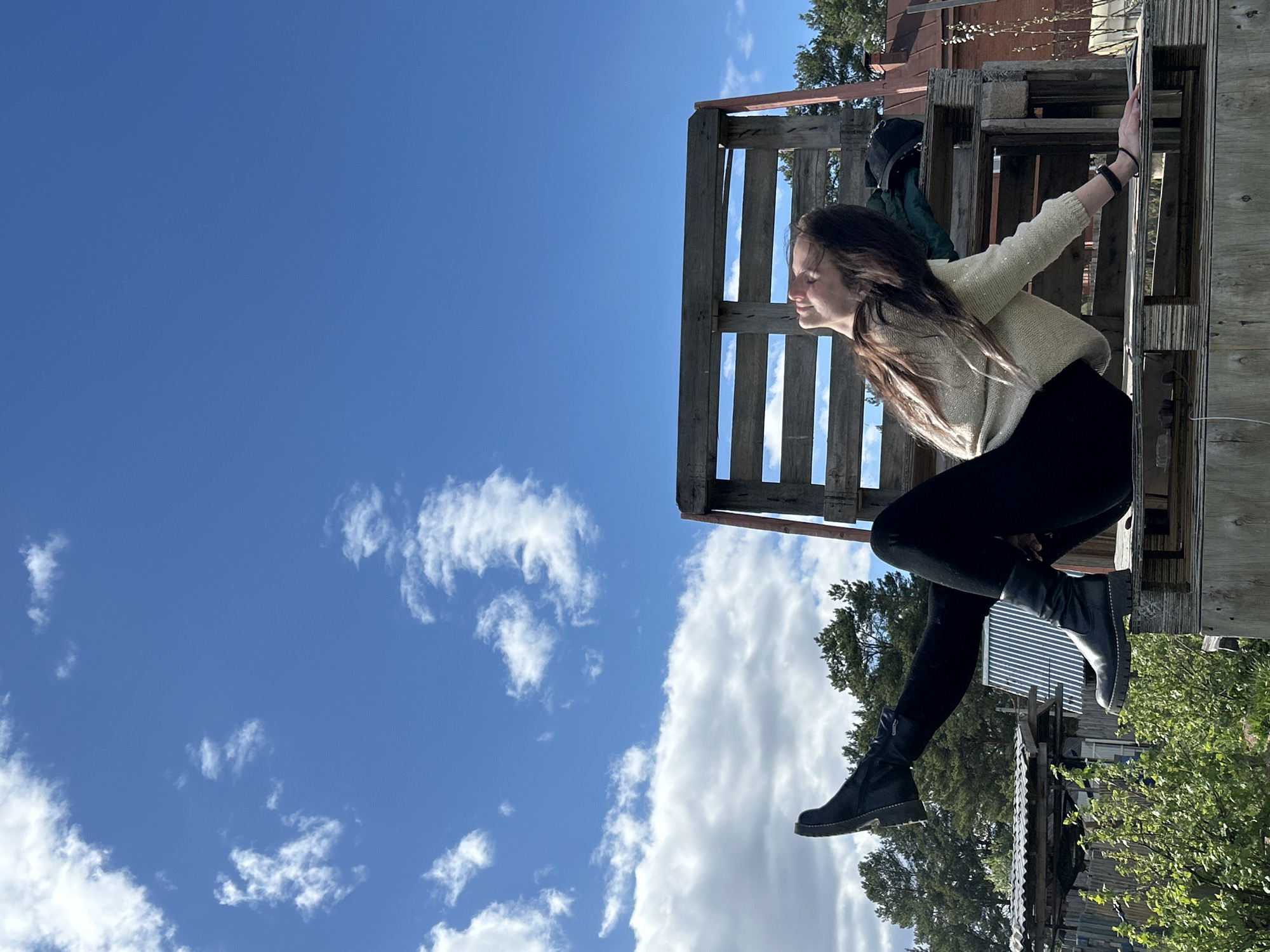 A woman is sitting on a wooden structure outdoors. The background is a blue sky with some clouds. The woman has brown hair and is wearing a light-colored sweater and black pants with black boots. She is looking to her left and smiling slightly. The wooden structure appears to be a pallet or part of a larger construction. Some foliage and parts of buildings are visible in the background. No evidence suggests the scene is in Rusalevka, Russia.
