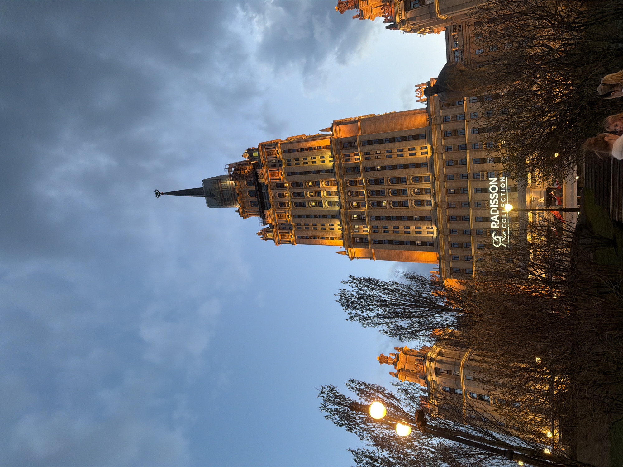 The image depicts the Radisson Collection Hotel, Moscow, a Stalinist skyscraper, viewed from below. The building is illuminated and contrasts against the evening sky. Bare trees are visible in the foreground, and street lights add to the ambiance. A Radisson sign is prominently displayed on the lower portion of the building. People are subtly present on the right side of the frame. The location is recognizable as part of the urban environment of Moscow, Russia.