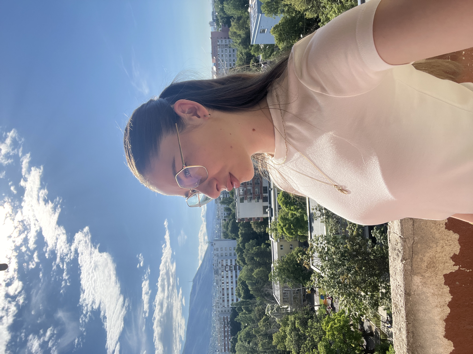 An outdoor, eye-level, medium shot shows a young woman standing on a balcony and looking to the right. She is wearing a white t-shirt and gold-rimmed glasses. The background shows a cityscape with buildings and trees, and mountains in the distance. The sky is blue with white clouds. The woman has long brown hair pulled back in a ponytail and is wearing a gold necklace with a pendant. The balcony railing is in the foreground.