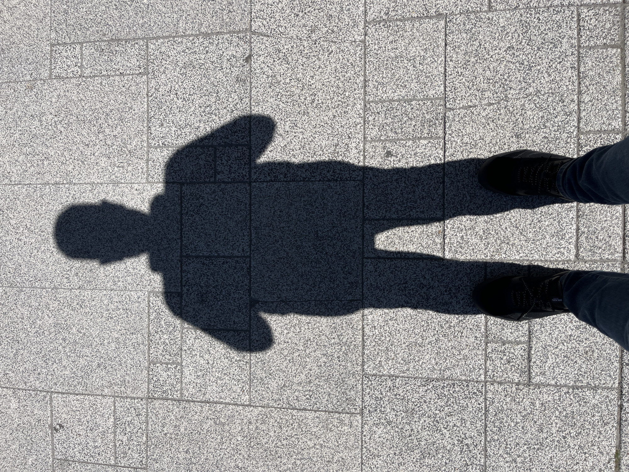 A full-body shadow of a person is cast on a granite tile sidewalk. The shadow is in the center of the frame, facing the camera. The person's head is at the top of the shadow, with arms bent at the elbows and hands near the waist. The shadow appears to be wearing a backpack. The feet of the person casting the shadow are visible at the bottom of the frame, wearing black sneakers and dark pants. The sun is bright, indicating it is daytime. The location is Unknown, Unknown.