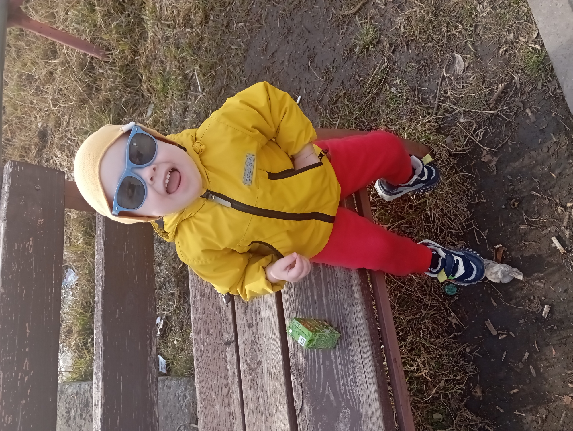 A young child sits on a park bench in Yurgamysh, Russia. The child wears a yellow jacket, red pants, a yellow hat, and blue sunglasses. They are smiling and looking towards the camera. A small green box sits next to them on the bench. The bench is made of weathered wood and is surrounded by dirt and grass. Cigarette butts are visible on the ground near the bench.
