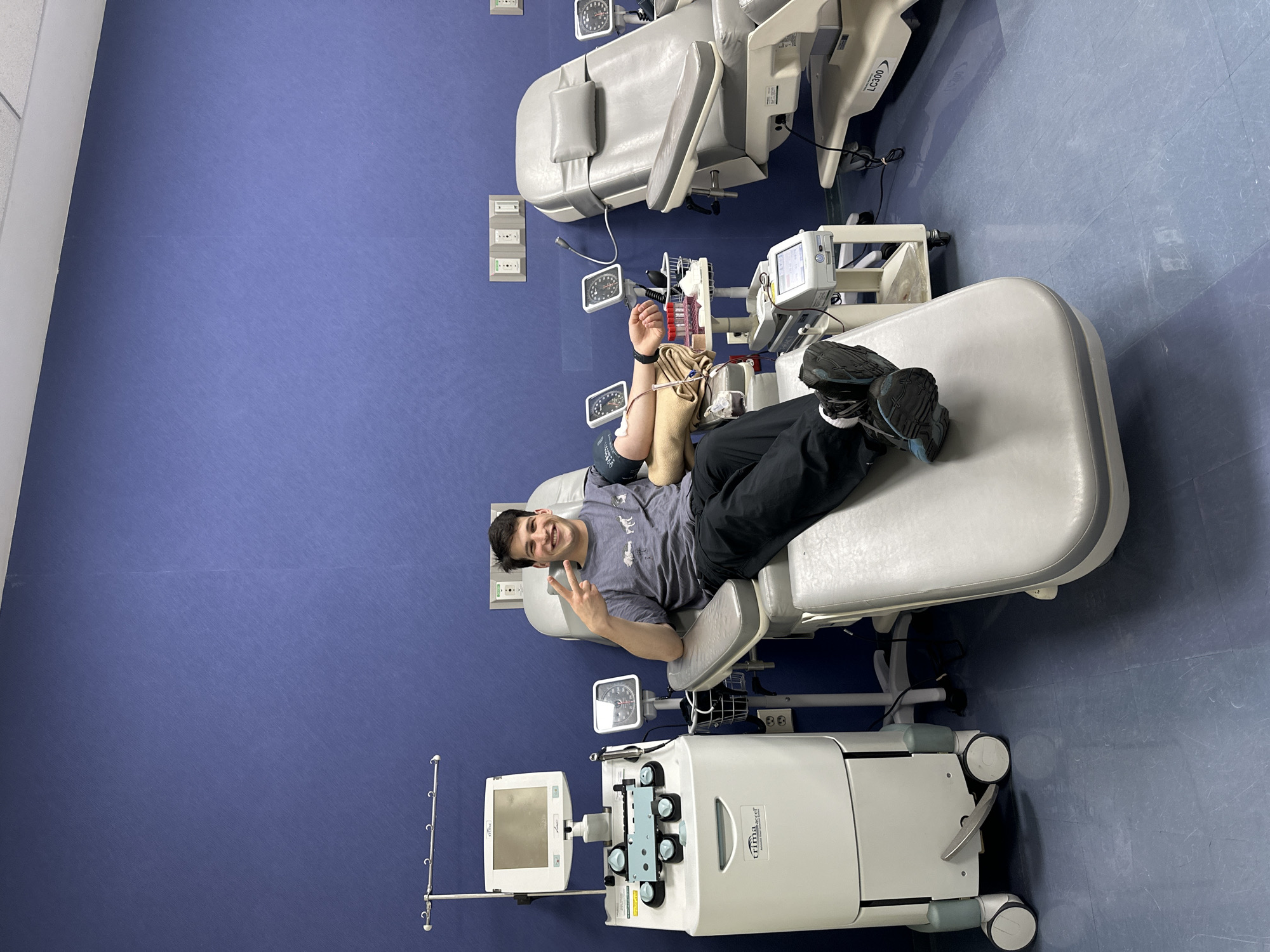 A young man is photographed in what appears to be a medical facility. He is reclining in a medical chair, connected to a blood donation machine, giving a peace sign. The room features medical equipment, a blue wall, and flooring. The location is indeterminate, but the city is identified as Huixquilucan de Degollado, Mexico.