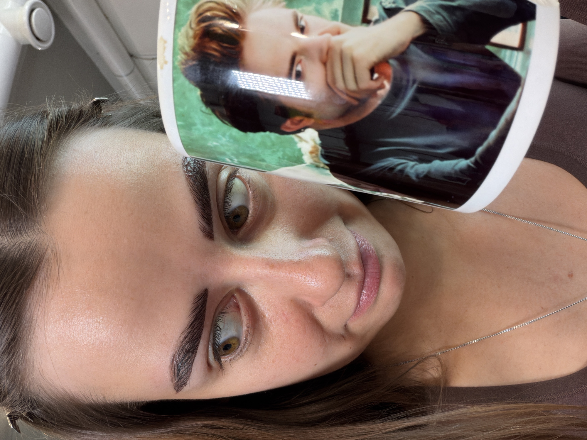 A close-up, angled shot shows a woman holding a mug with a printed image. The woman's face is partially visible, with her eyes looking to the left. She has dark, thick eyebrows and her lips are slightly parted. She is wearing a brown shirt and a silver necklace. The mug features a picture of a man with blonde-highlighted hair, wearing a dark shirt and resting his hand over his mouth. The background of the image is out of focus but appears to be an indoor setting, possibly an office or home. There is no visible indication of the specific location within Krasnoyarsk, Russia.