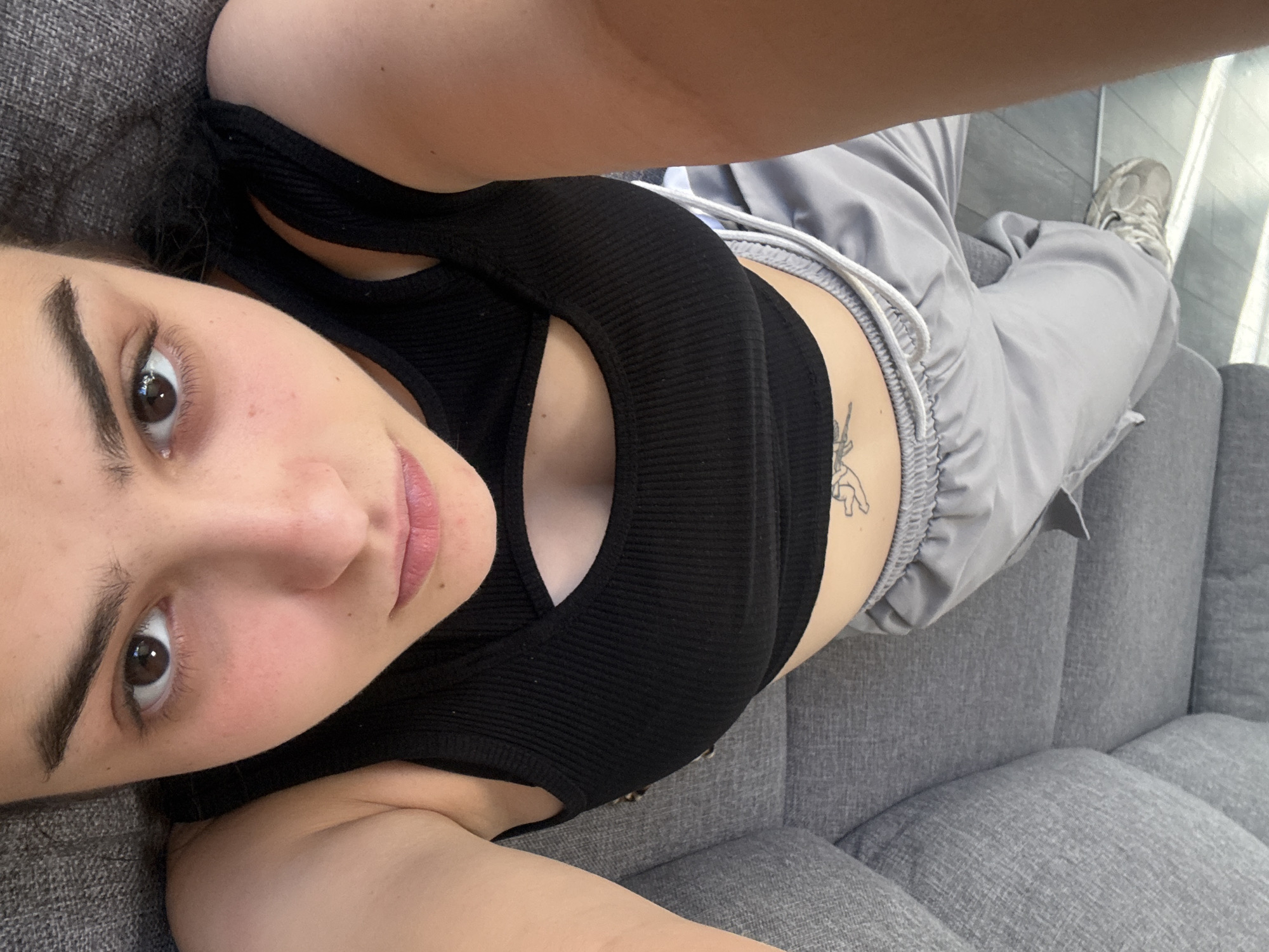 A woman is lying on a gray sofa. She is wearing a black tank top and gray sweatpants. She has a tattoo on her abdomen. Her right leg is stretched towards the right of the photo. She has brown hair and eyes. The photo is taken from above. The exact location in Blagoevgrad, Bulgaria, cannot be determined from the image.