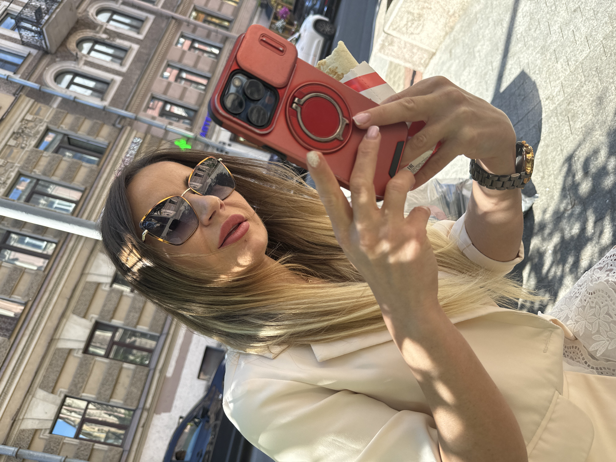 The media file shows a close-up, upward-angled selfie of a woman on a sunny day. The woman, with blonde hair and wearing sunglasses and a cream-colored jacket, is holding a red smartphone with a protective case. The phone case has a circular ring stand. The woman's right hand is visible, wearing a watch with a dark band and a gold-toned face. She is also holding a partially eaten sandwich in a red and white wrapper. The background shows a building with multiple windows, suggesting an urban setting. The sunlight casts shadows, indicating it is likely daytime. The overall impression is of a casual moment captured outdoors. Some text is visible on the sandwich wrapper, reading "EAC" in capital letters. There is also some faint text on the phone case.