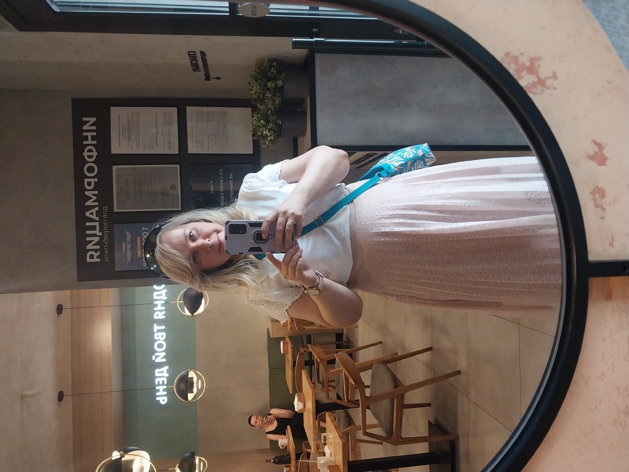 Here's a matter-of-fact description of the image:
A woman with blonde hair is taking a selfie in a round mirror, likely inside a cafe or restaurant. She is wearing a white blouse and a light pink, pleated skirt. A turquoise handbag strap is visible across her shoulder. The mirror reflects the cafe interior, including tables, chairs, and a person seated at one of the tables. Decorative hanging lamps and signage with the Russian text "RNHAМΠOΦΗΝ" and ""заждүднэдо эондод" are also visible. The OCR also identifies "CLINORG", "HE NOT RHAC", "100", "DAH Y", "CO CBOEN ETON", "OTRING H".
Based on the signs, the location is Orenburg, Russia.