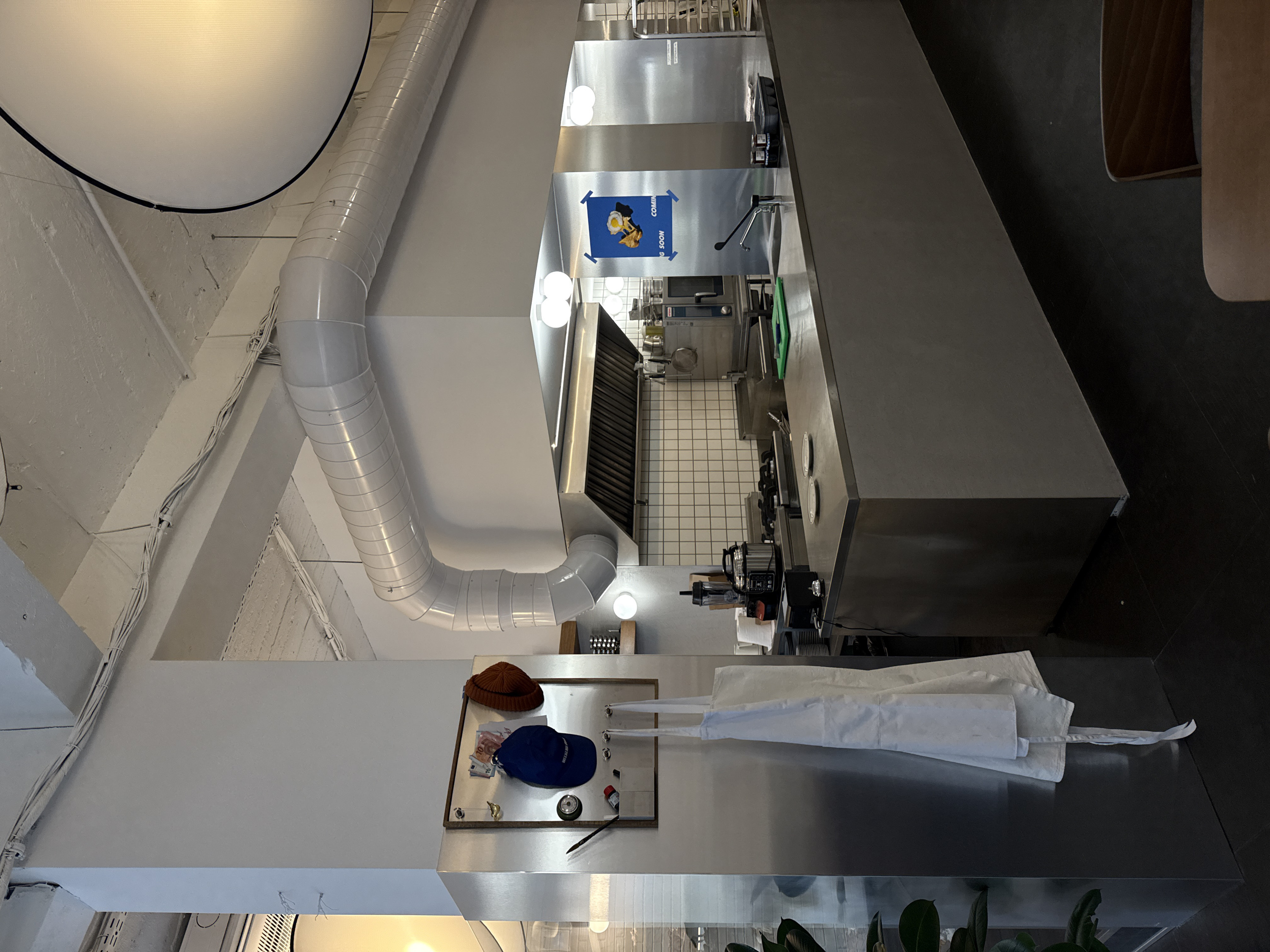 This image captures an interior view of JOSH, a street food and breakfast establishment located at ul. "Georgi S. Rakovski" 108, 1000 Tsentar, Sofia, Bulgaria. The scene depicts an unoccupied, brightly lit professional kitchen and prep area with a modern industrial aesthetic.
The environment features a white-painted concrete ceiling with exposed white spiral-wound air ducts and electrical conduits running along beams. Walls are predominantly white, with a white square-tiled backsplash behind the main cooking station. The floor is dark.
A large, grey stainless steel island counter dominates the midground. On its left, a black receipt printer, a small silver service bell, a black blender, and a silver multi-cooker are positioned. Centrally, two empty white ceramic plates rest on the counter. To the right, a green plastic cutting board, a chrome sink faucet with a black handle, and a black tray containing several small glass jars are arranged.
Behind the island, a commercial gas range with multiple burners, a large stainless steel extractor hood, and a deep fryer are visible. Stainless steel panels form partitions in the kitchen area. A blue poster featuring a fried egg dish with "COMING SOON" and "JOSH" branding is taped to one of these panels.
On the far left, a tall, brushed stainless steel clad column displays a wooden-framed magnetic board. Attached items include two Euro banknotes (10 and 20), a blue baseball cap with "WEEKEND CLUB" lettering, an orange knit beanie, a small gold bird ornament, a round green gauge, and a black pen. A white chef's apron hangs from hooks below the board. A large potted plant with dark green leaves is partially visible at the bottom left.
Multiple spherical white wall sconces illuminate the kitchen area, complemented by a large, white, suspended spherical light fixture visible in the upper right quadrant. A wooden chair back and a portion of a wooden tabletop are visible at the bottom right, suggesting an adjacent dining area. No individuals are present in the image.